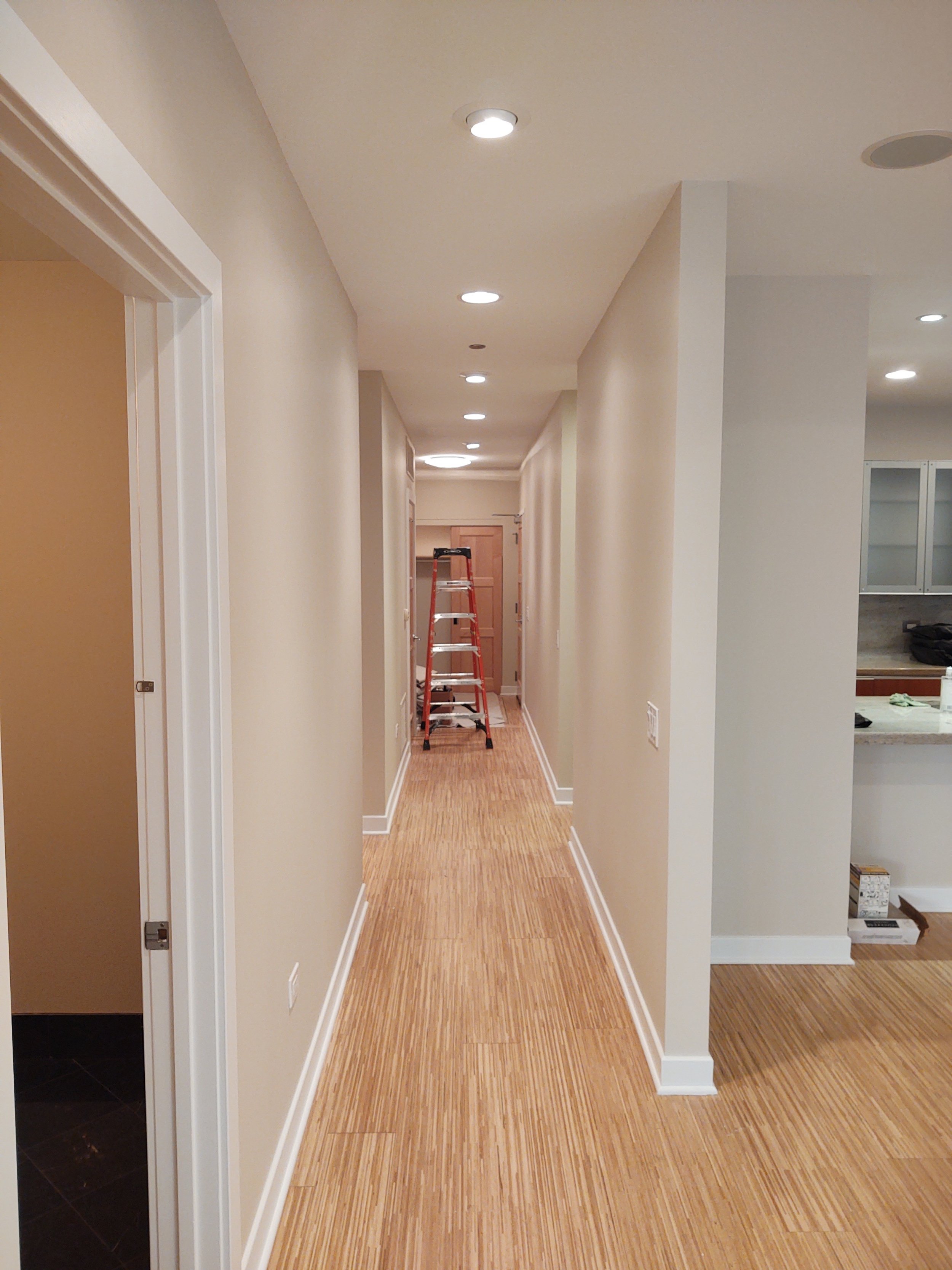 Empty hallway with recent renovation, beige walls, wood flooring, recessed ceiling lights, ladder in the distance, kitchen visible on the right.