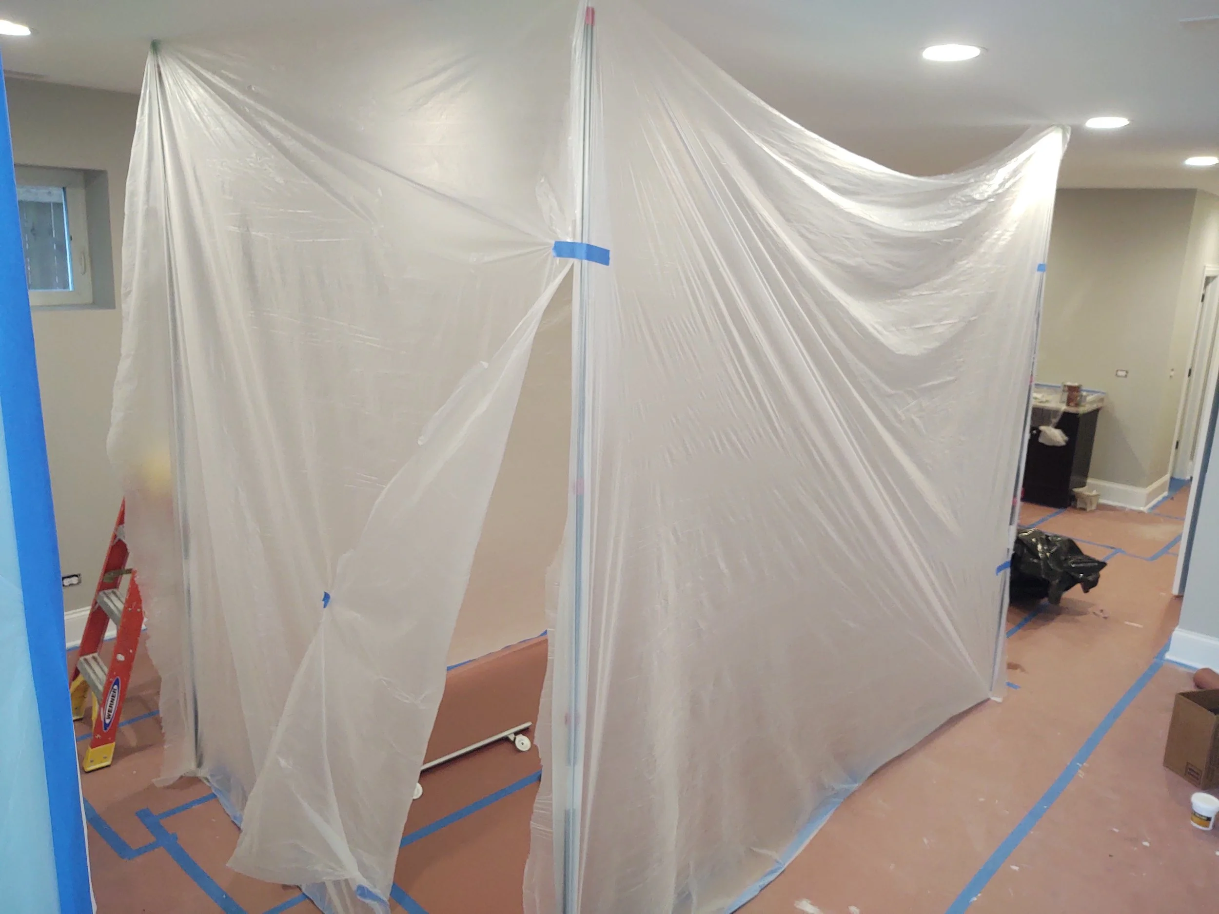 A room under renovation with plastic sheeting covering walls or furniture, a ladder on the left, and painter's supplies on the floor and in the background.