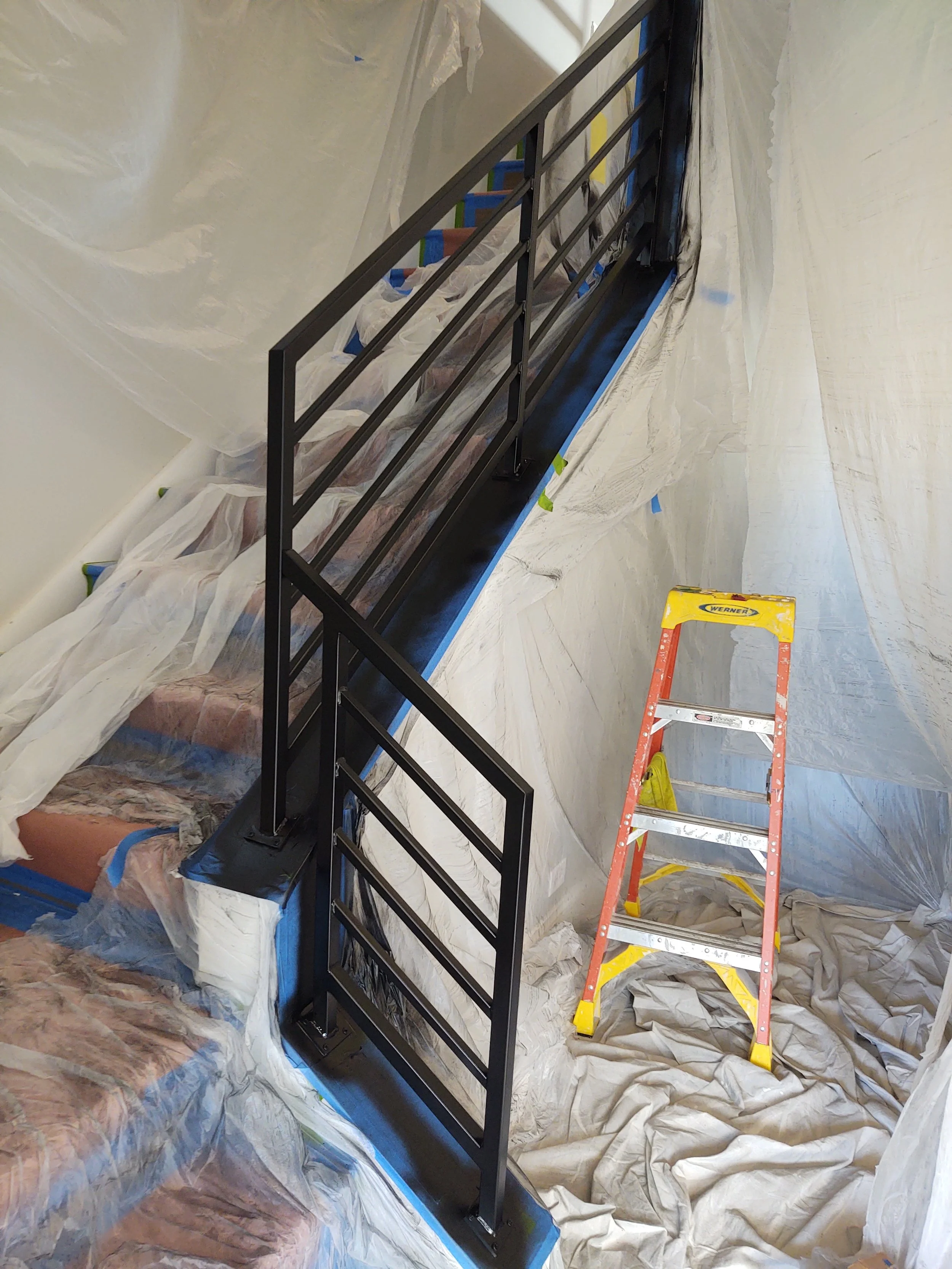 Black metal railing being installed next to a staircase in a construction site, with a yellow and orange step ladder nearby and plastic sheeting covering the surrounding area.