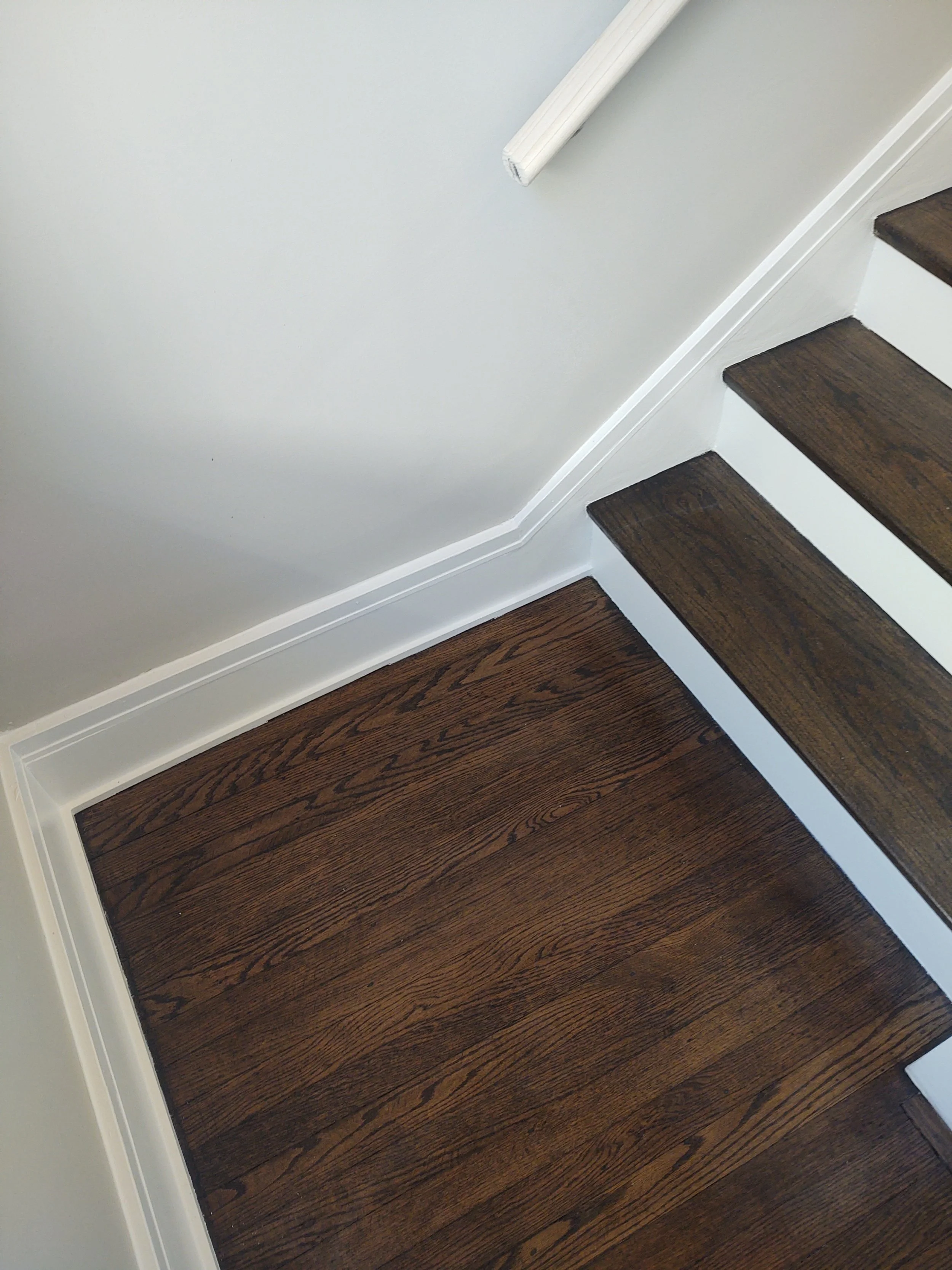 Close-up of a corner of a finished staircase with dark wooden treads, white risers, and white wall with baseboard.