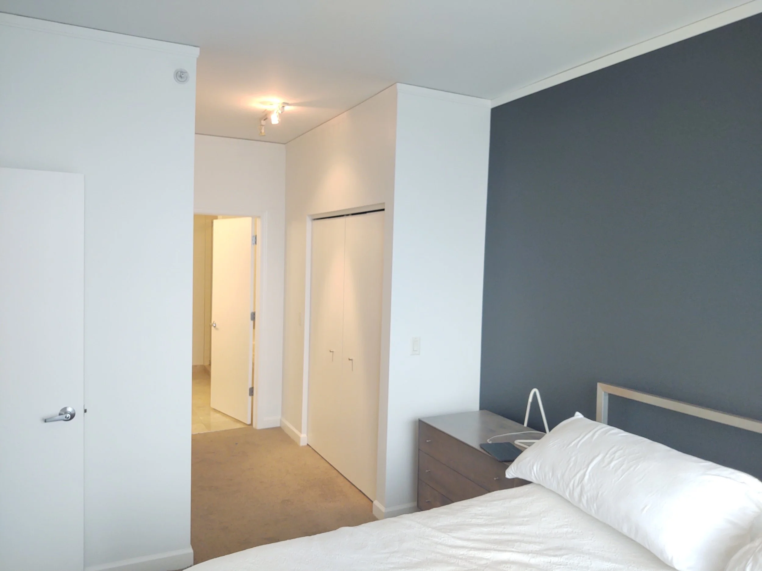A bedroom with a bed featuring white bedding and a pillow, a gray two-drawer nightstand with a laptop and reading lamp, white walls with a dark accent wall, a closet with sliding doors, and a doorway leading to a brightly lit room or hallway.