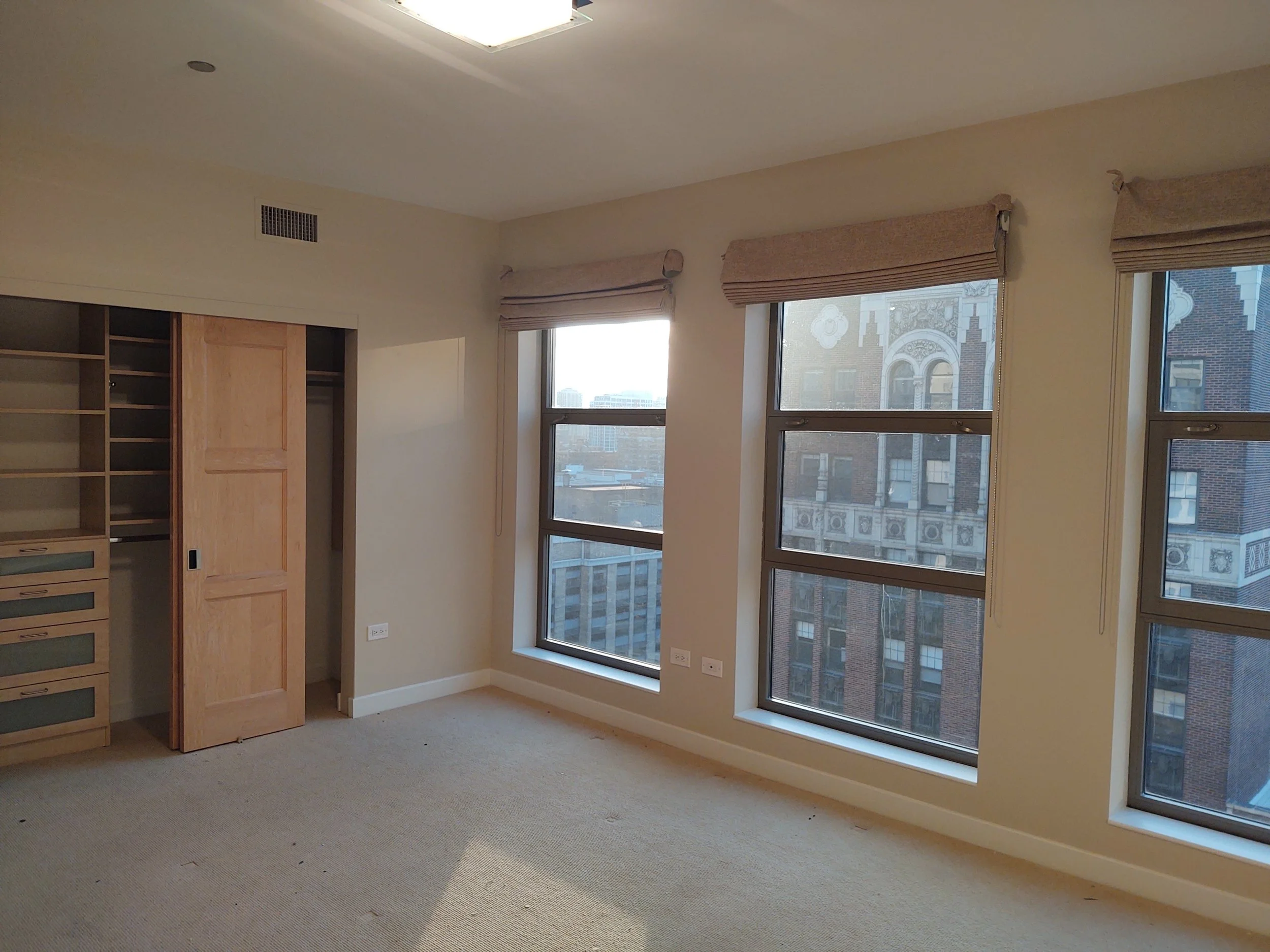 Empty room with beige carpet, three large windows with beige valances, built-in wooden closet with open shelves and drawers, and city buildings visible through the windows.