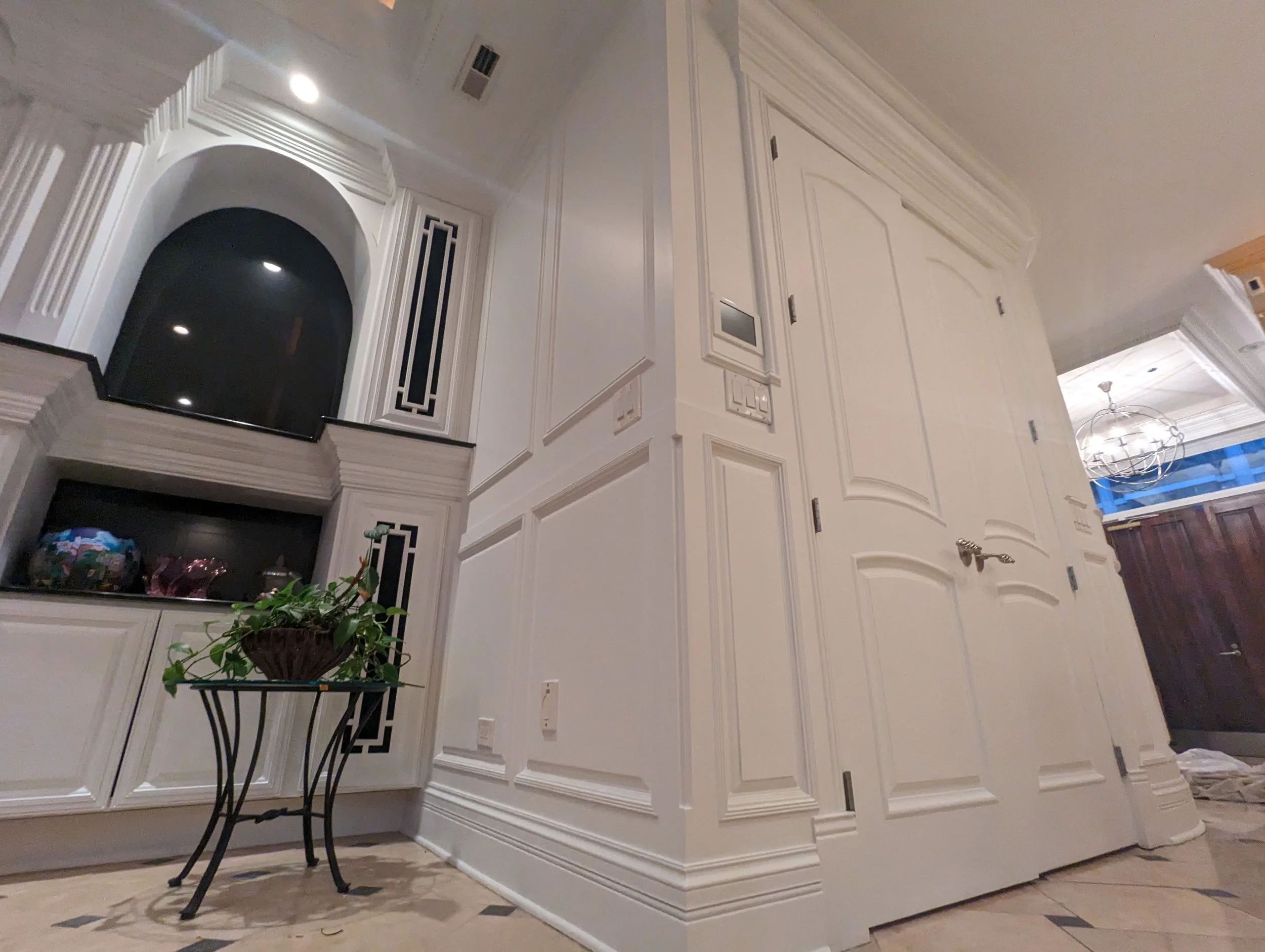 Interior of a luxurious home with white detailed walls, a closed white door with a silver handle, a small black table with a potted plant, and decorative black and white cabinetry. Visible part of a dark wooden door, and a chandelier through the door