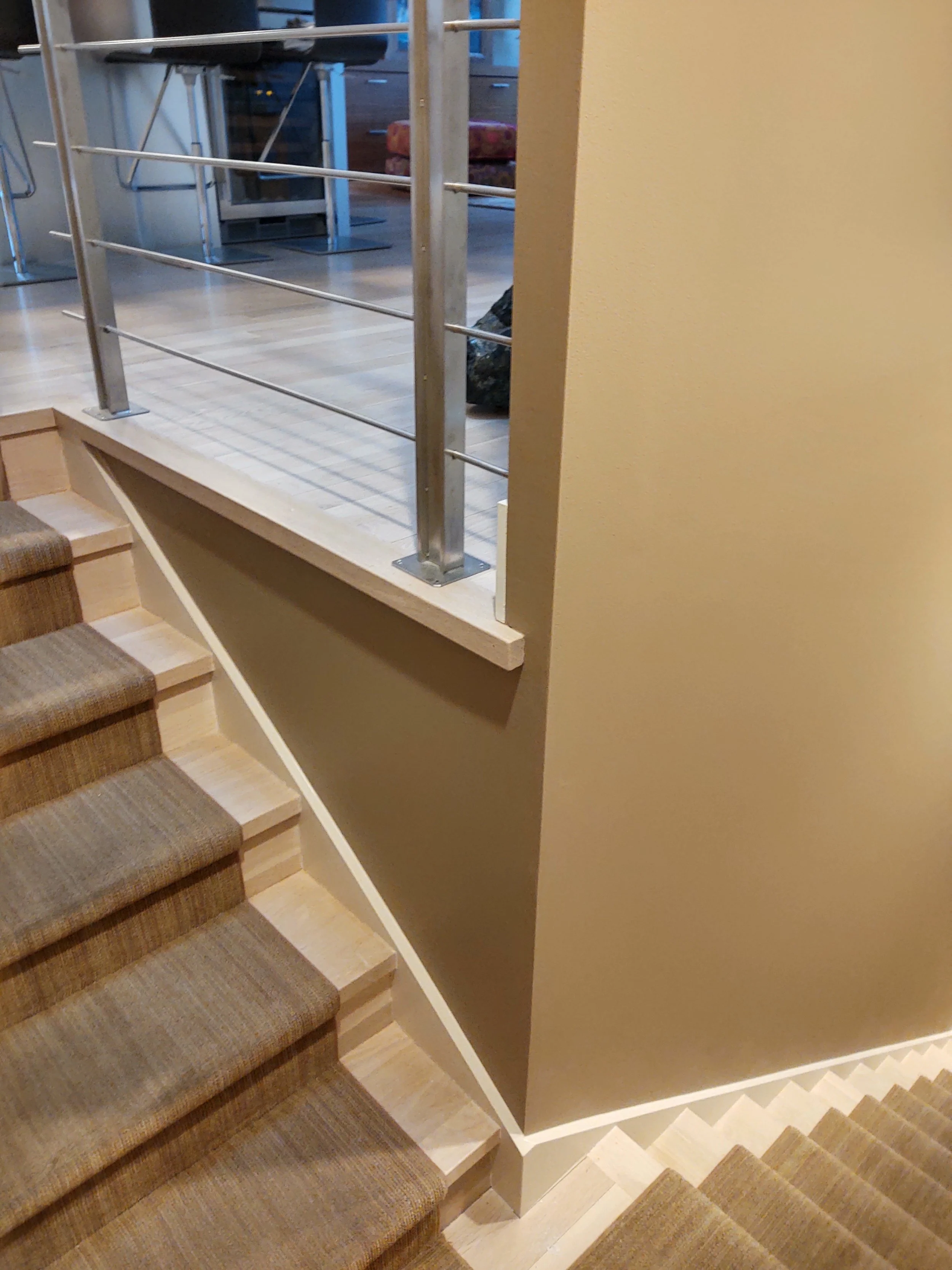 Photo of a staircase with brown carpeted steps, a beige wall on the left, and a beige wall with a white baseboard on the right. A metal handrail with horizontal bars runs along the top of the staircase, attached to a partial wall on top of the steps.