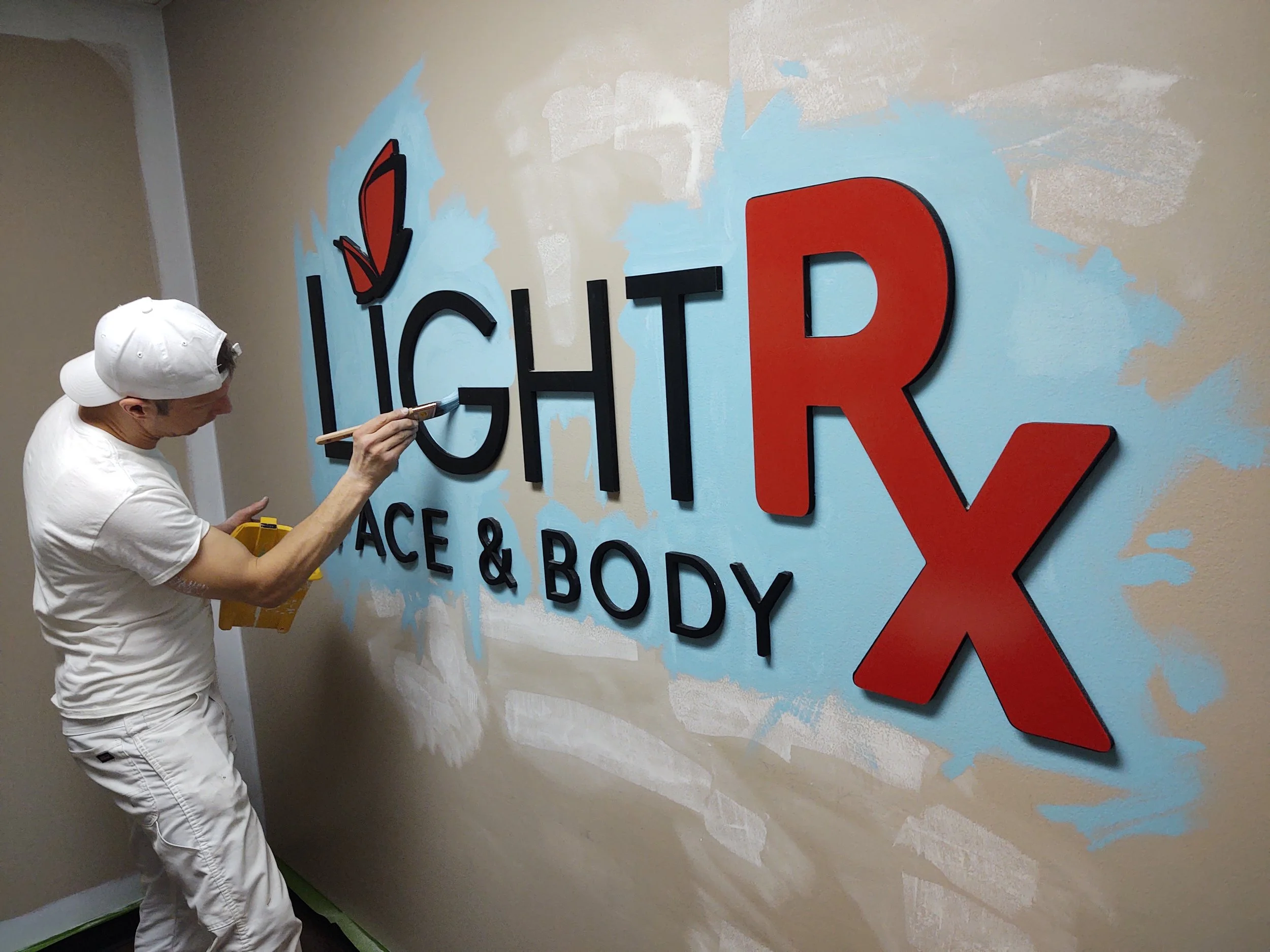 A person in white clothes and a white cap is painting a wall with a logo that reads 'LIGHT R FACE & BODY' with a red butterfly above the 'i' in 'LIGHT'. The wall has a light blue background with some unfinished patches.