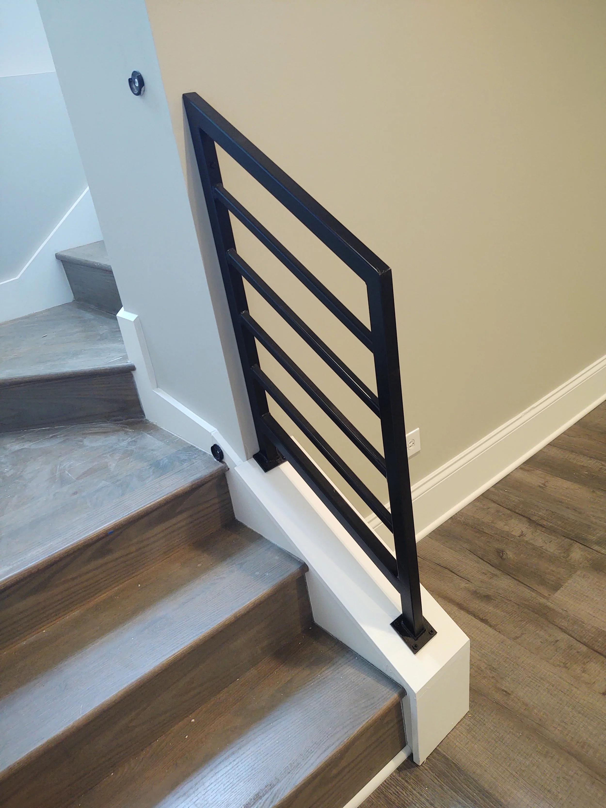 Photo of a staircase with a black metal railing on the right side, connected to a beige wall, with wooden steps.