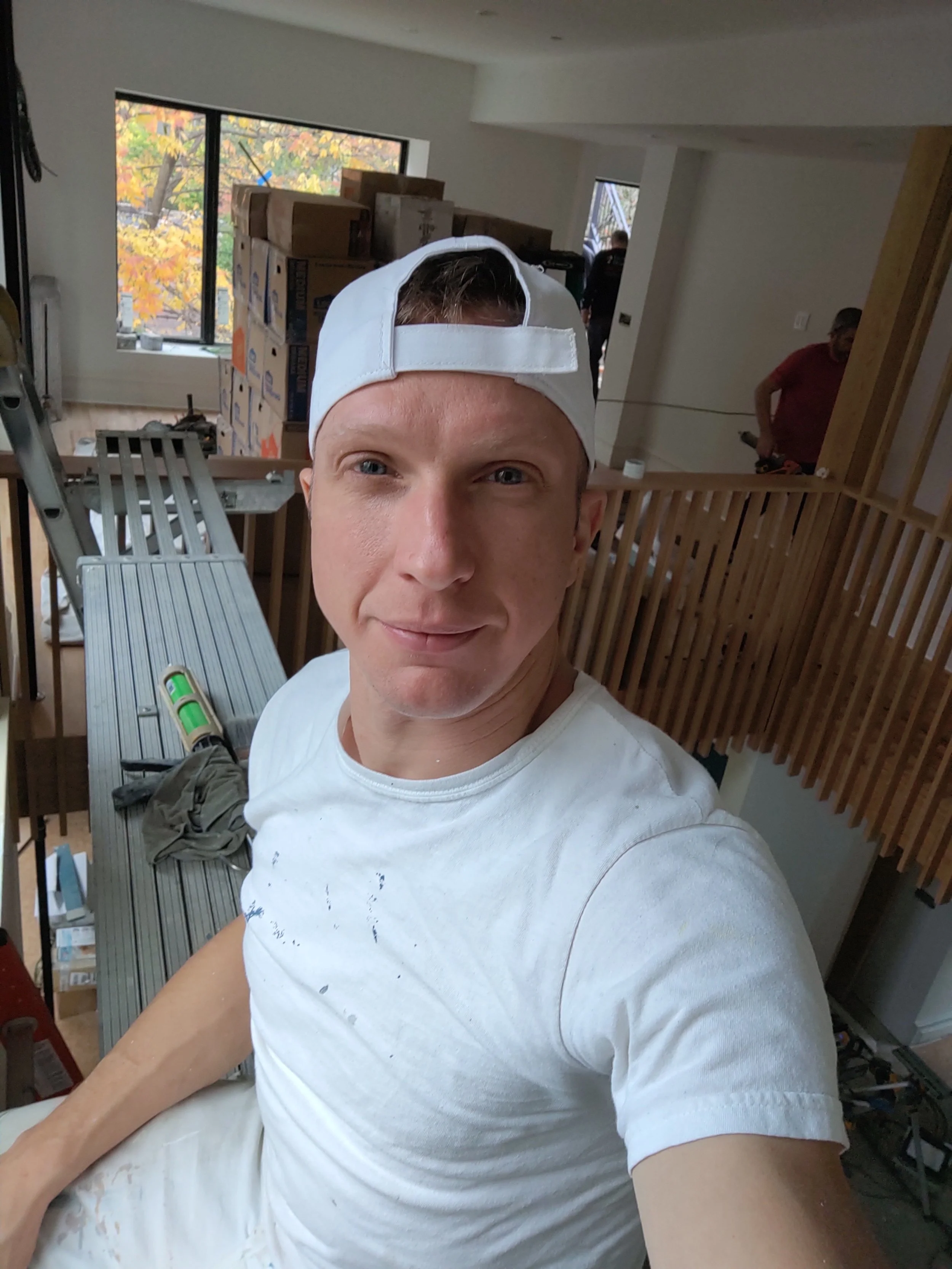 A young man wearing a white baseball cap backwards and a white t-shirt taking a selfie indoors. Behind him are boxes, a ladder, and various tools, indicating ongoing construction or renovation work.