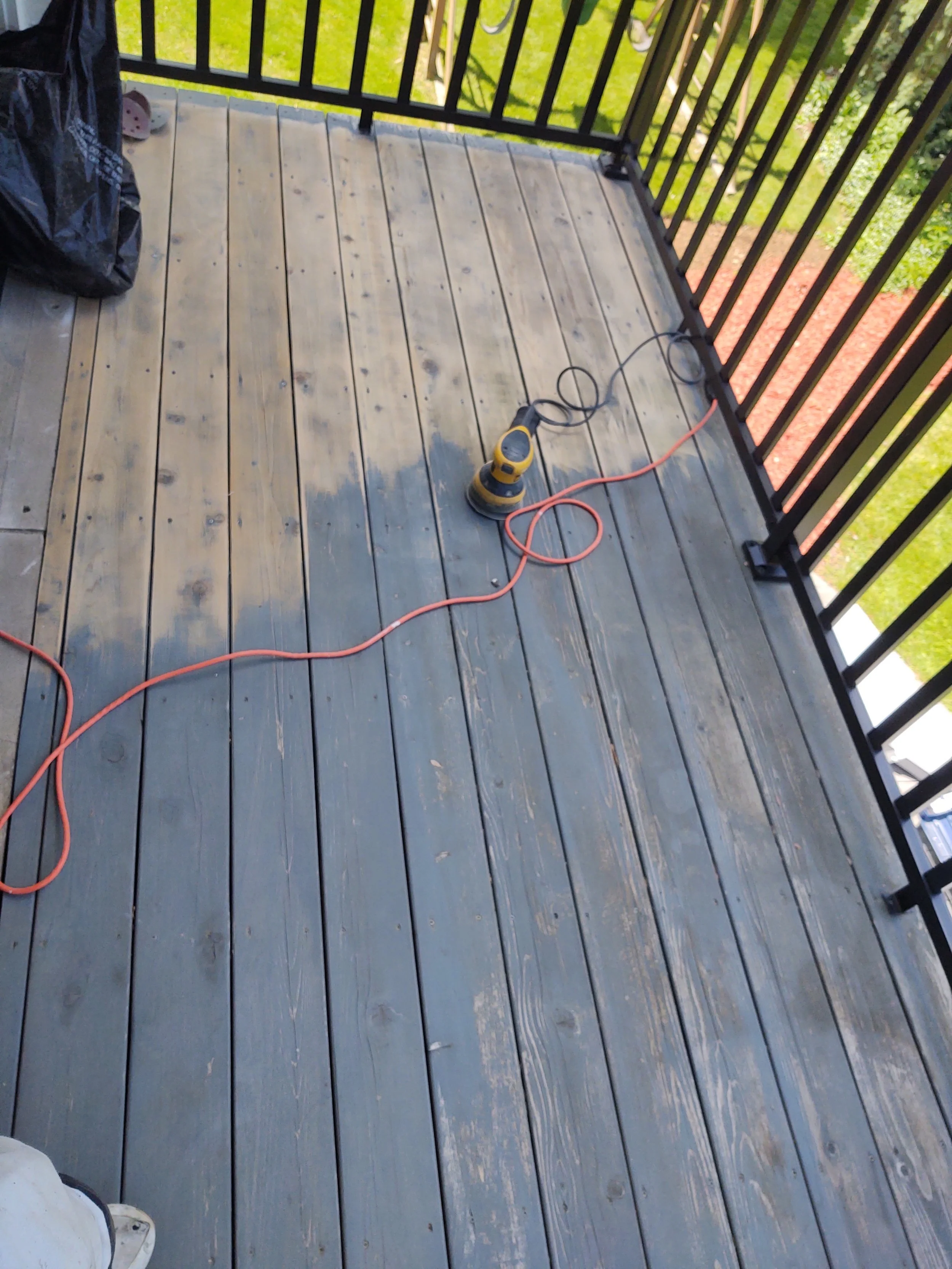 A wooden balcony with black railing looking down at an orange extension cord and a yellow portable power sander on the floor.