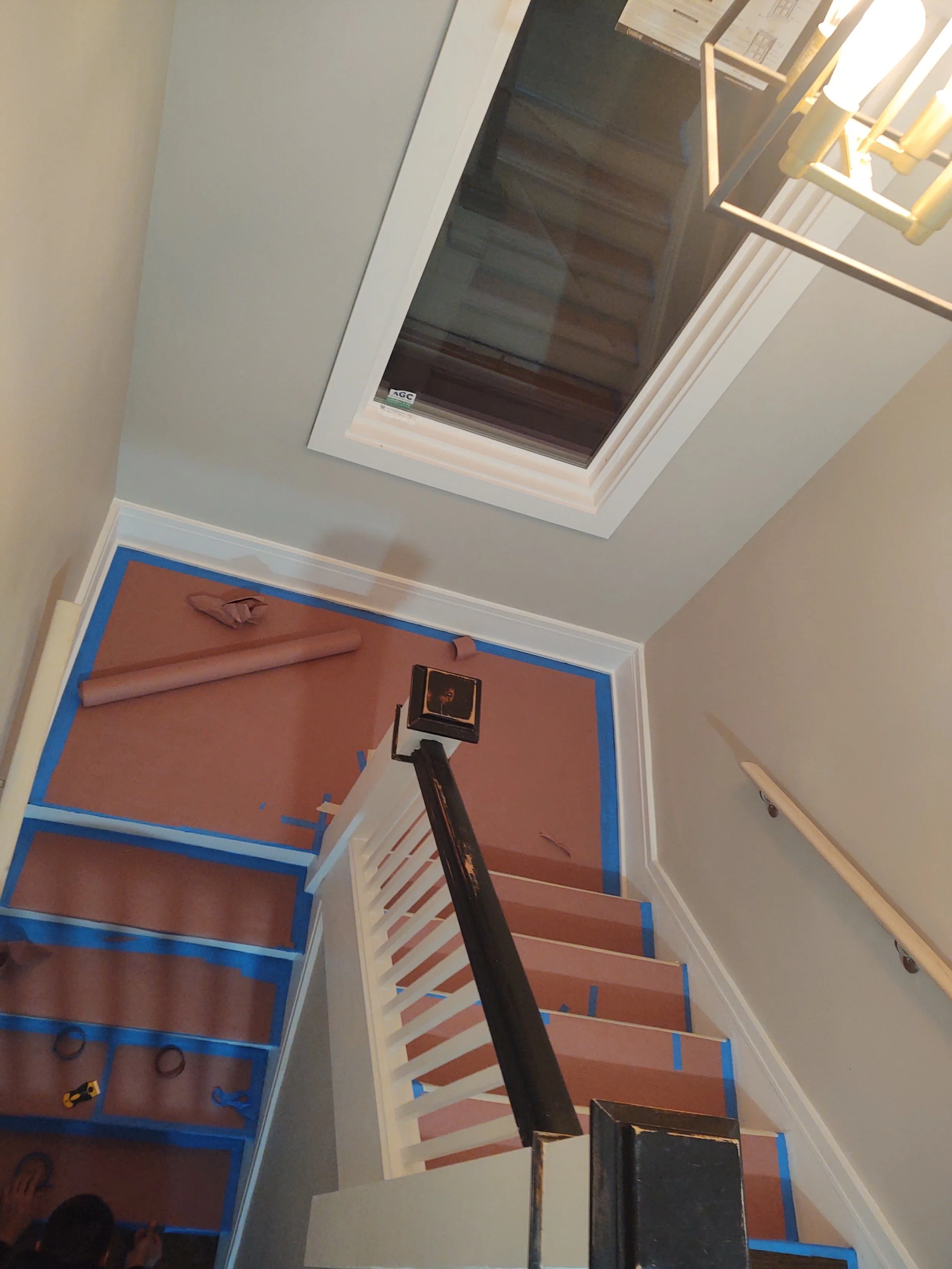 Inside a house, staging or renovating a staircase area with brown paper covering the carpet on the stairs. Blue painter's tape is visible along the edges of the stairs and wall. A light fixture hangs from the ceiling. A window is above the staircase,