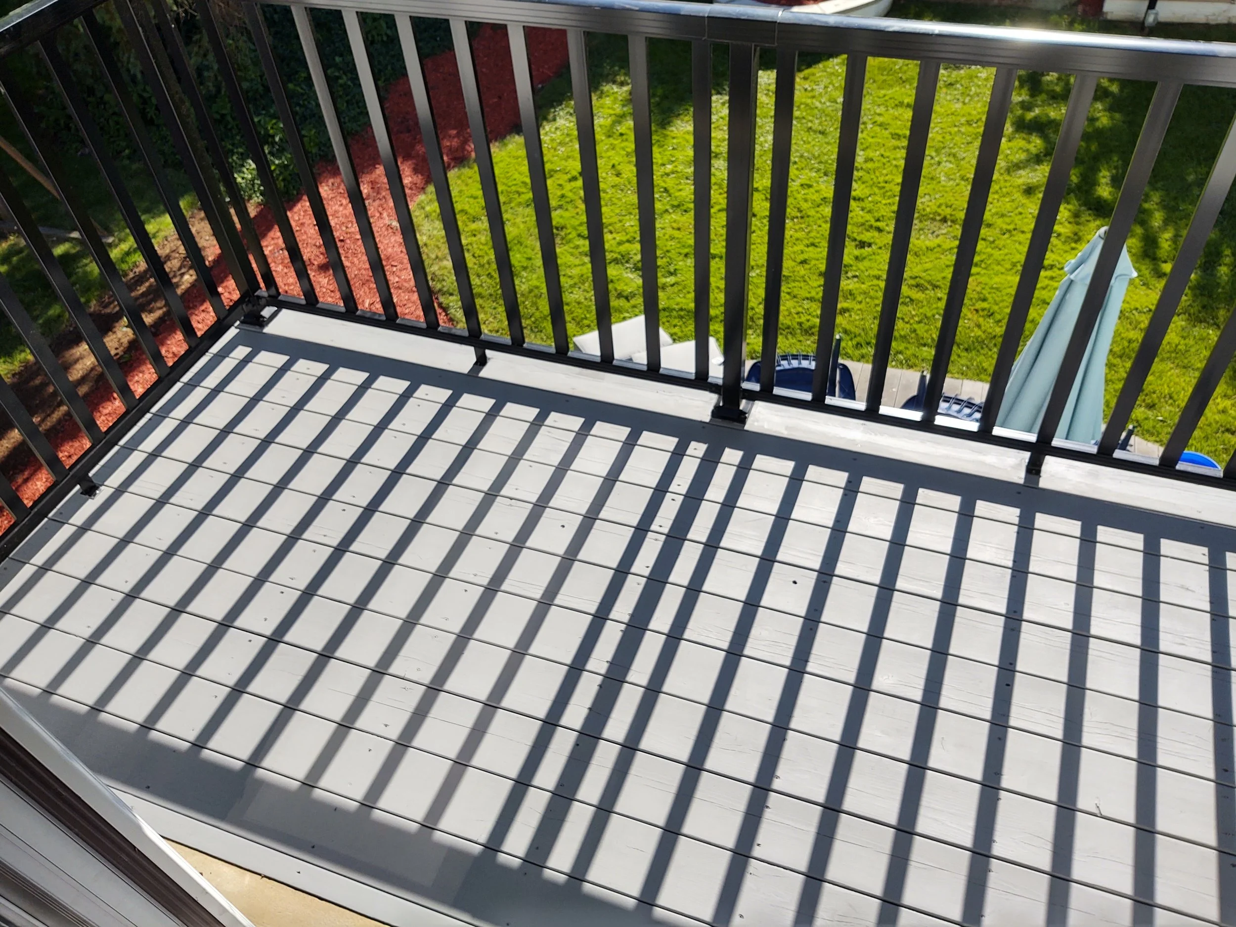 Small outdoor balcony with white wooden floorboards and black metal railing, casting striped shadows on the floor. Green grass, a garden bed with red mulch, and a patio with outdoor furniture and a closed umbrella are visible below.