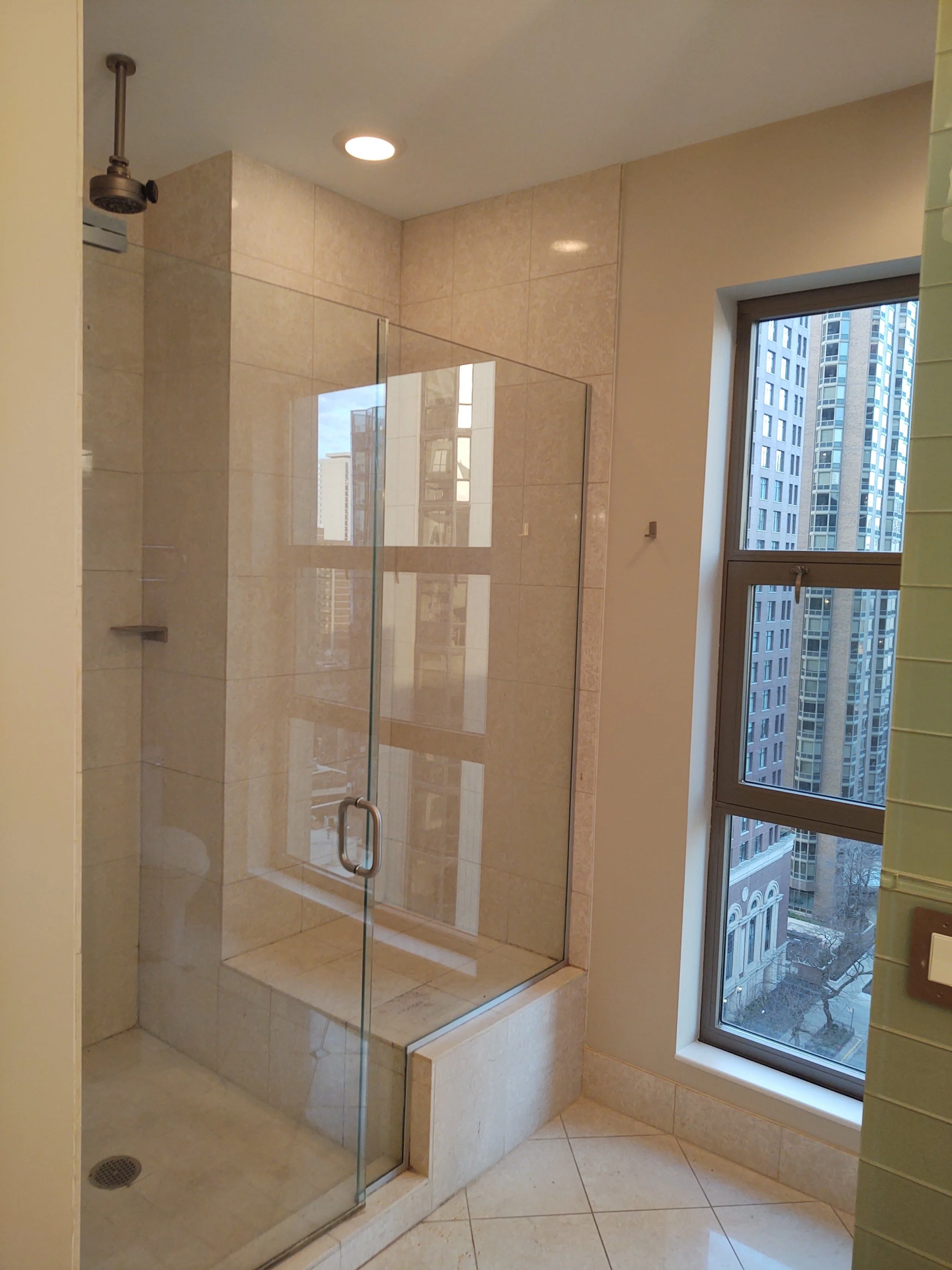 A shower with glass door, beige tiled walls, and a window showing high-rise buildings outside.