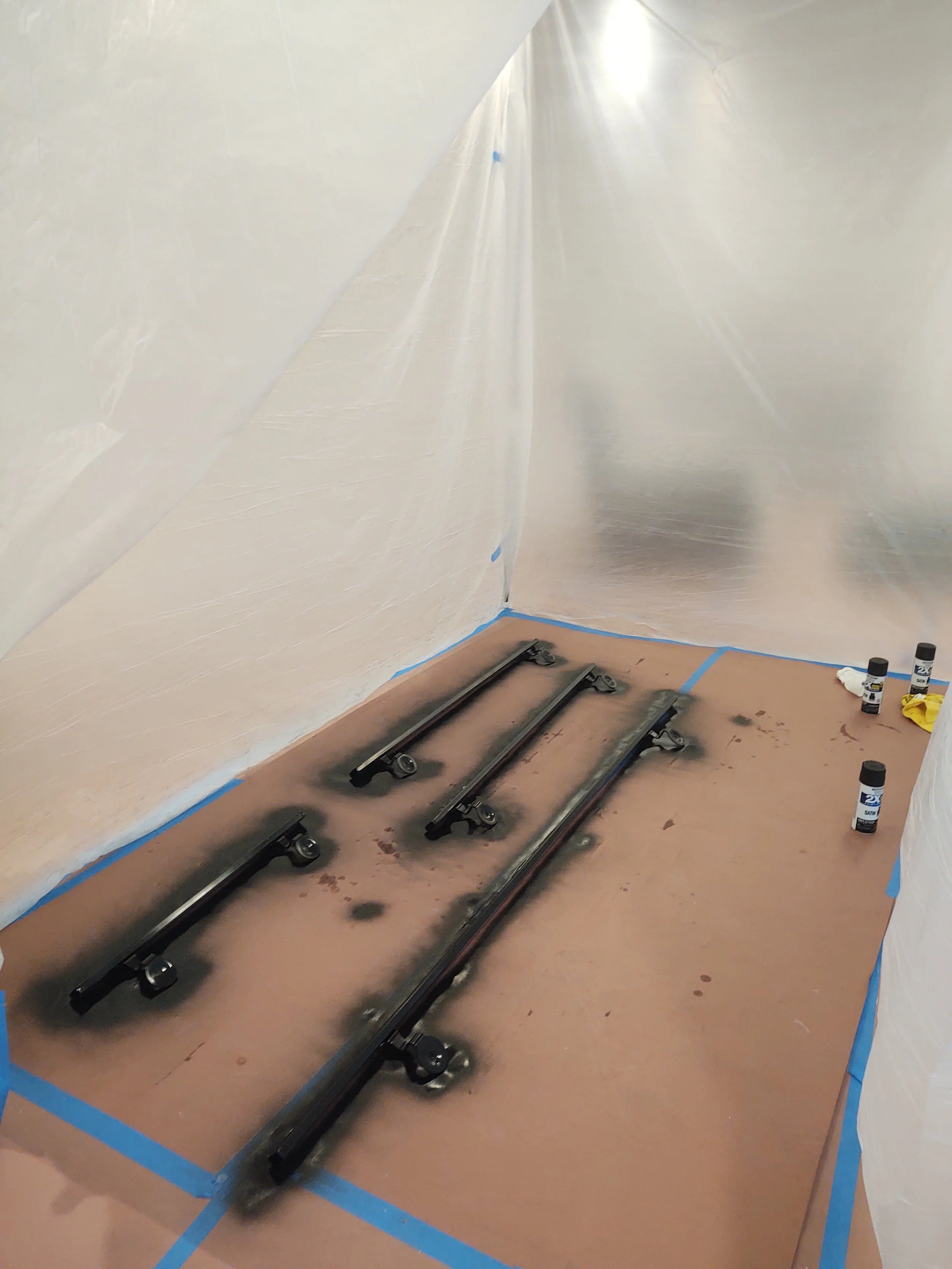 Three black spray-painted rails placed on a brown paper-covered floor in a room with white plastic sheeting walls, with spray paint cans and cleaning supplies nearby.