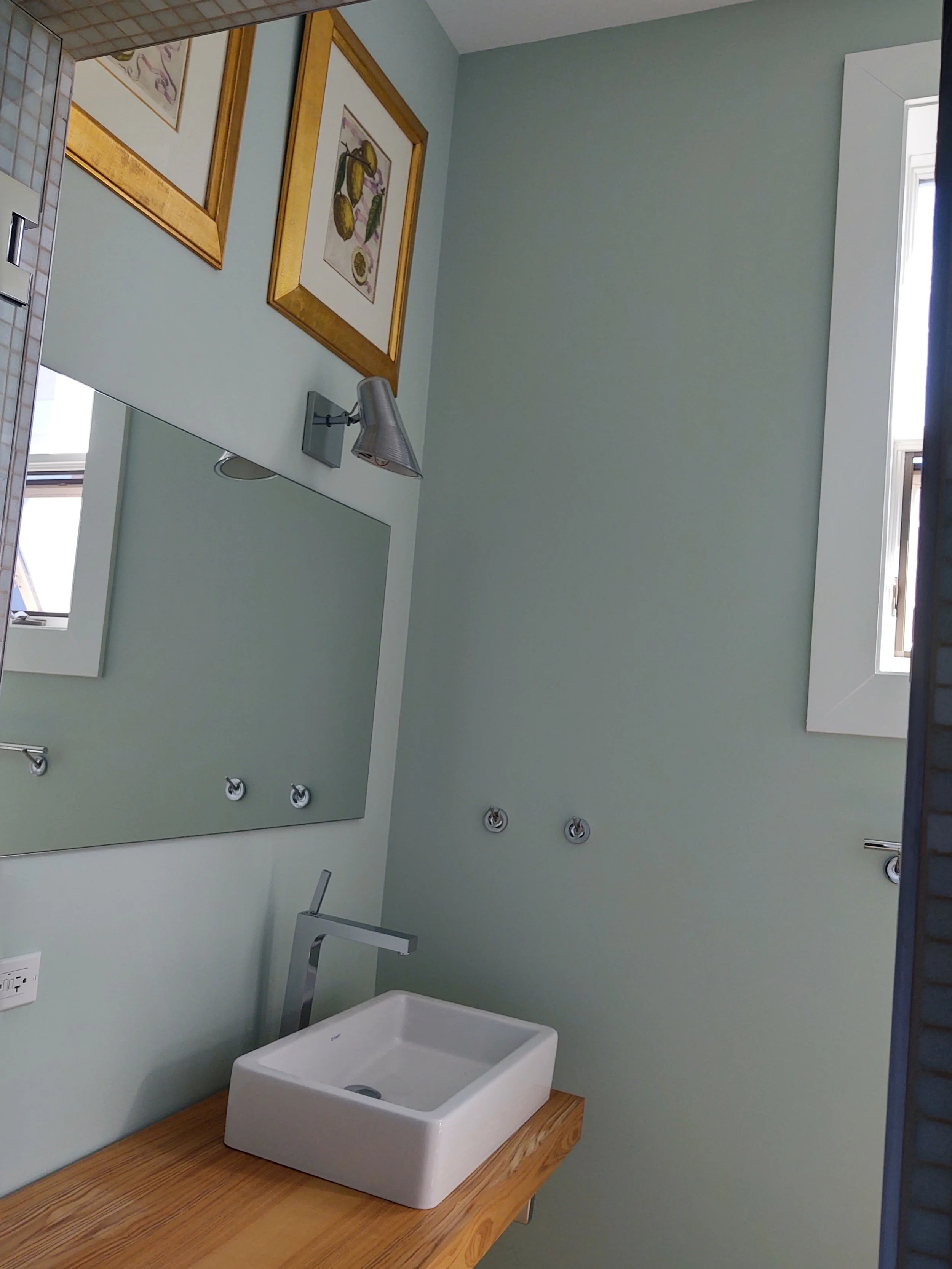 A bathroom with a rectangular white sink on a wooden countertop, a large mirror, two framed pictures above the mirror, a wall-mounted light fixture, and a small window with open shutters.