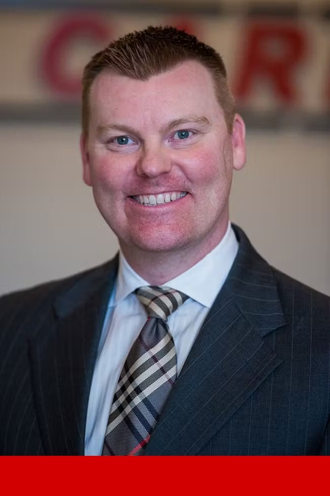 A man with short red hair, blue eyes, and light skin, smiling in a suit and tie, standing in front of a blurred background with red lettering.