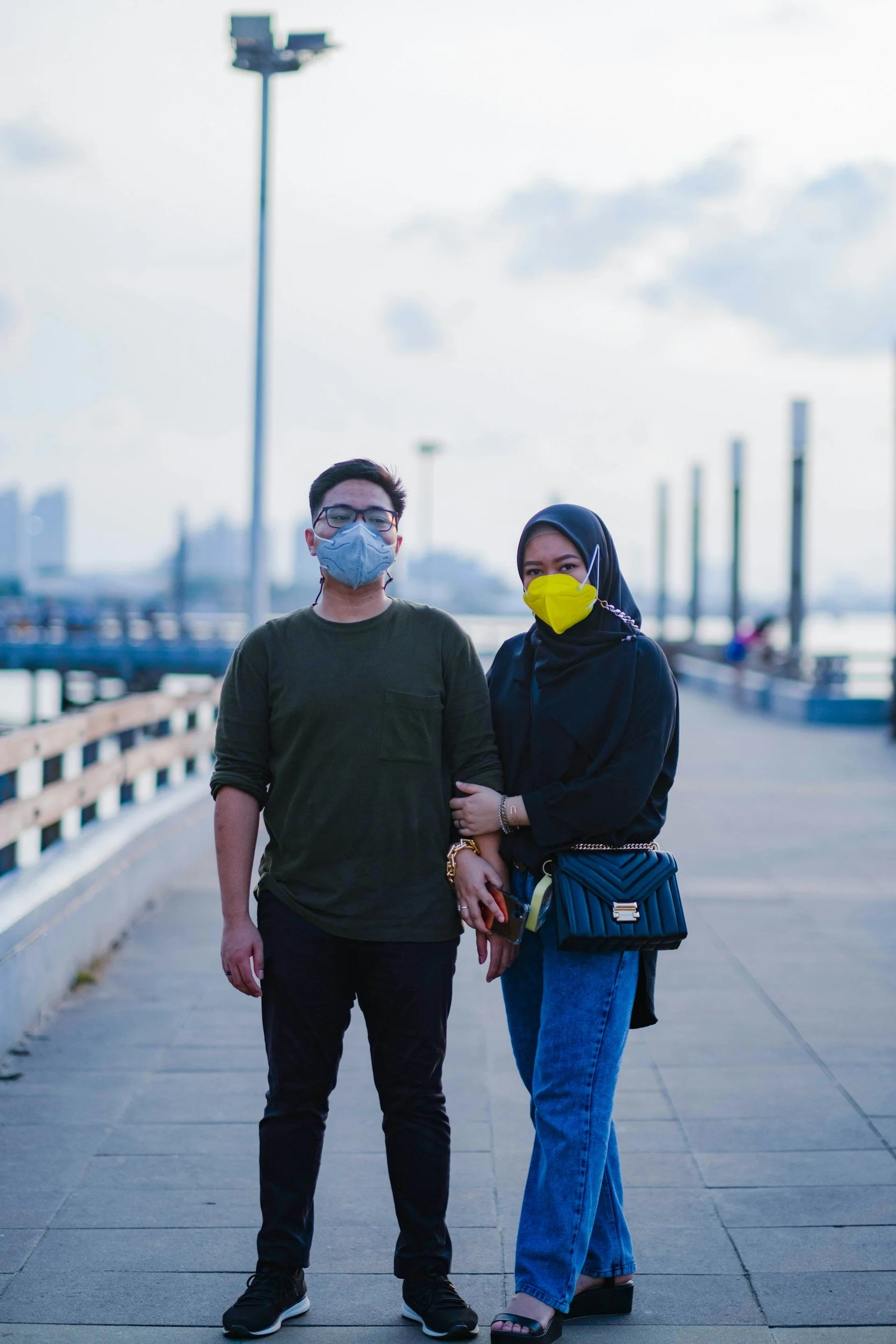 An Asian man with short dark hair wearing black glasses, a blue N95, and a green longsleeve tee and dark jeans and a Muslim woman wearing a black longsleve shirt, black hijab, yellow KN95, black purse, and jeans, stand arm and arm on a pier.