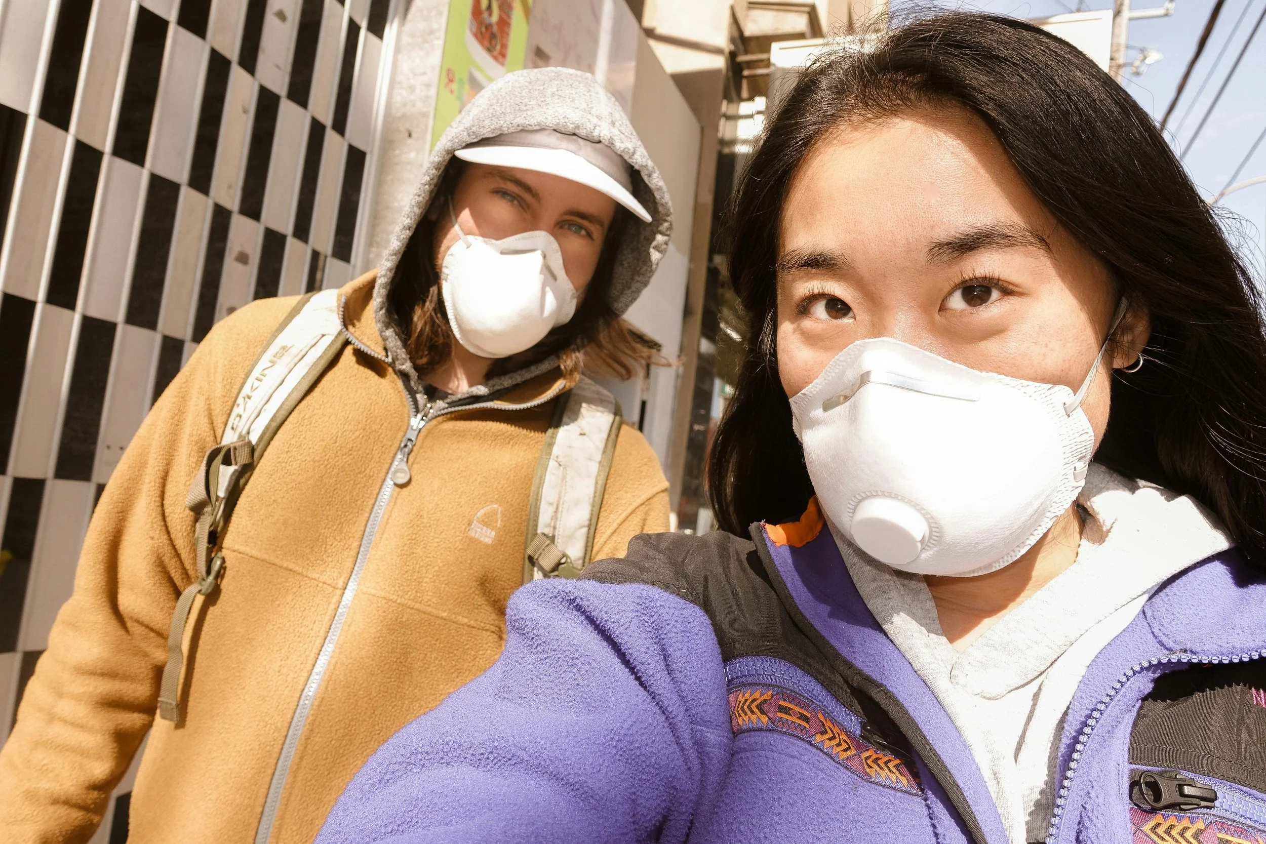 A white person in a taupe hoodie and an N95 takes a walking selfie in the city with an Asian woman wearing a vented N95 and purple coat.