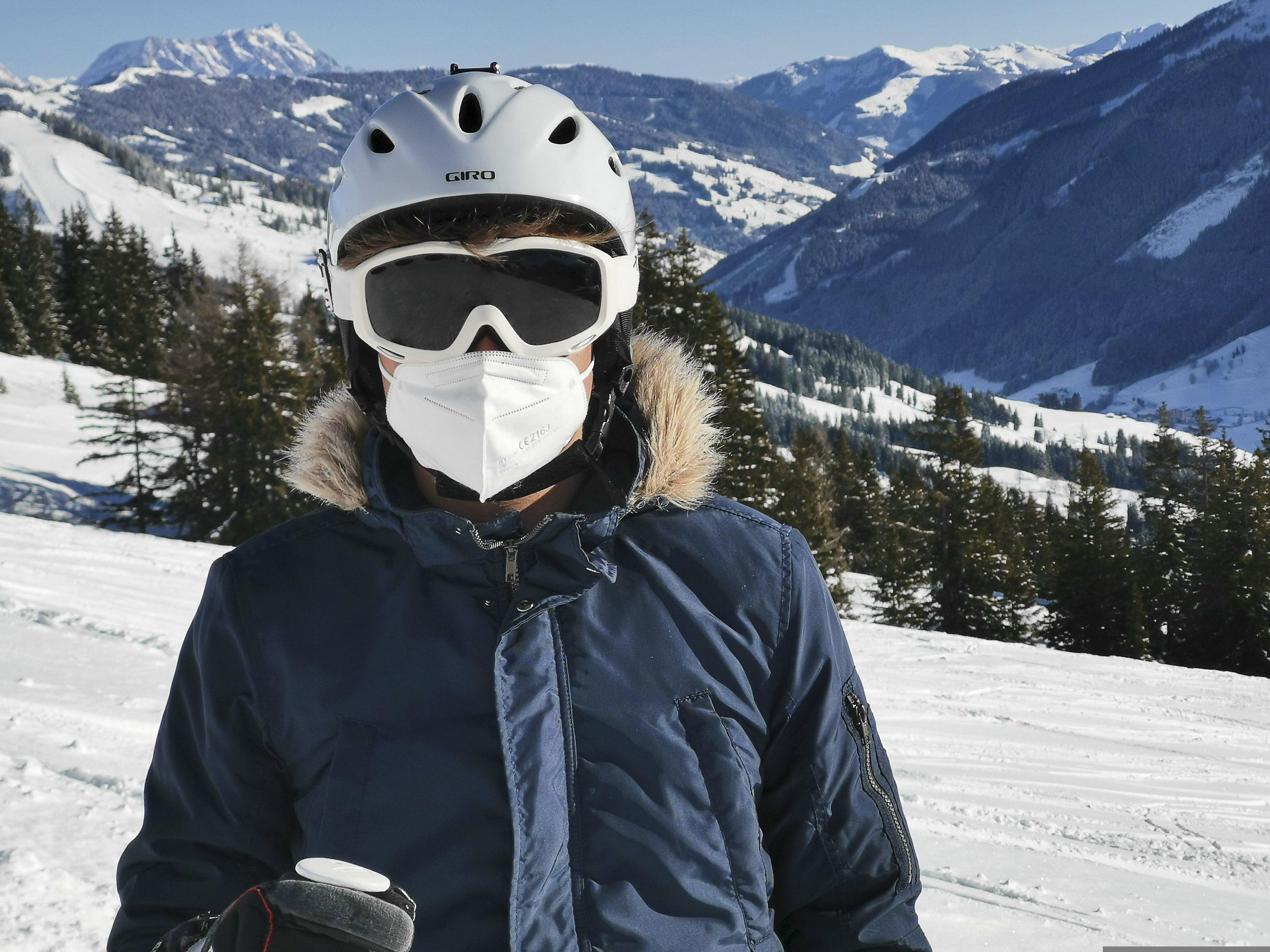 Person in navy blue ski jacket with brown fur collar, silver ski helment, white goggles, and a white N95 stands on a snow-covered mountain with some evergreen trees in the background.