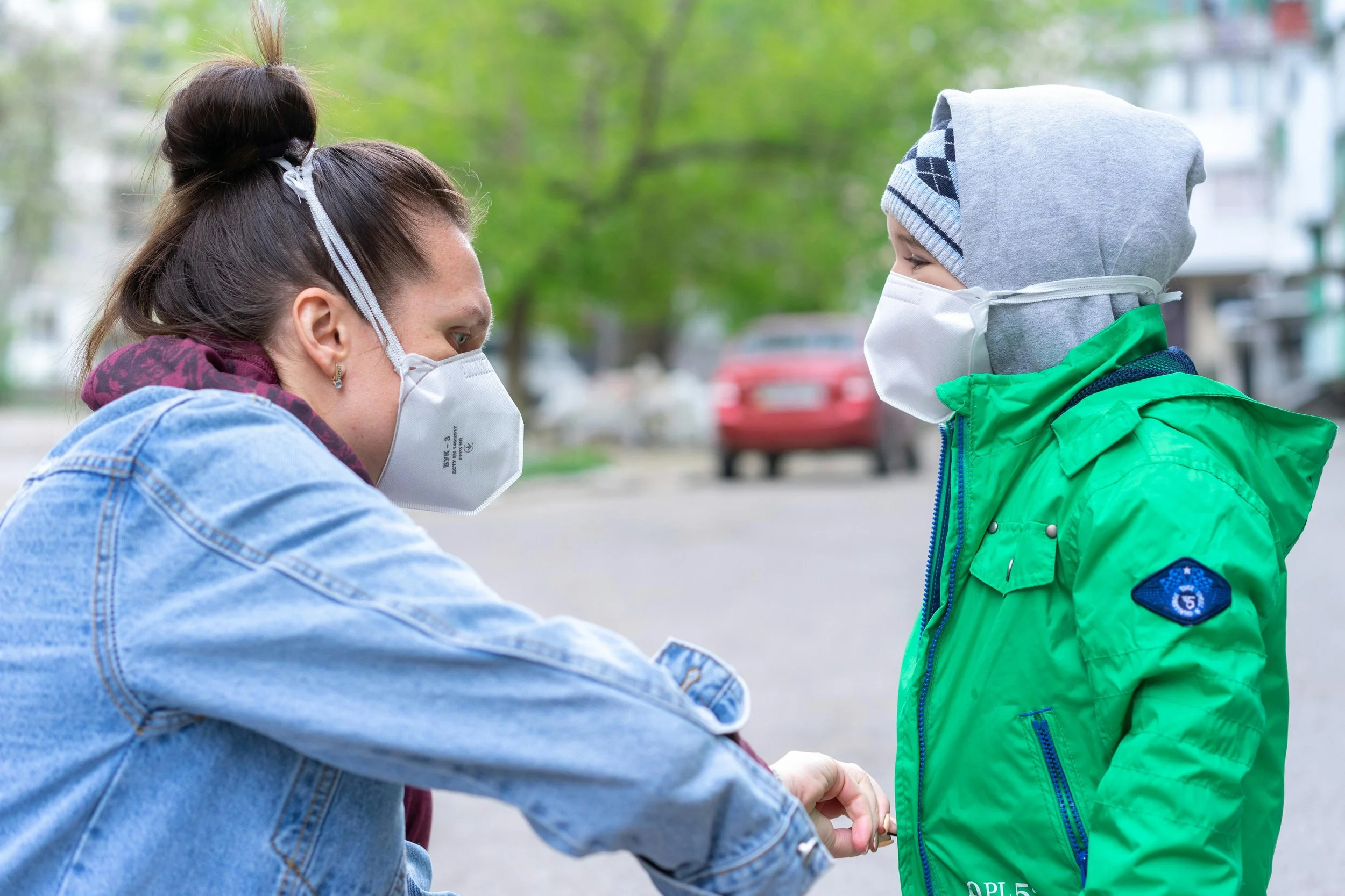 A middle-aged brunette white woman in a blue denim jacket and N95 helps a young white boy in a lime green jacket over a grey sweatshirt with the hood up and an N95 zip up his coat.