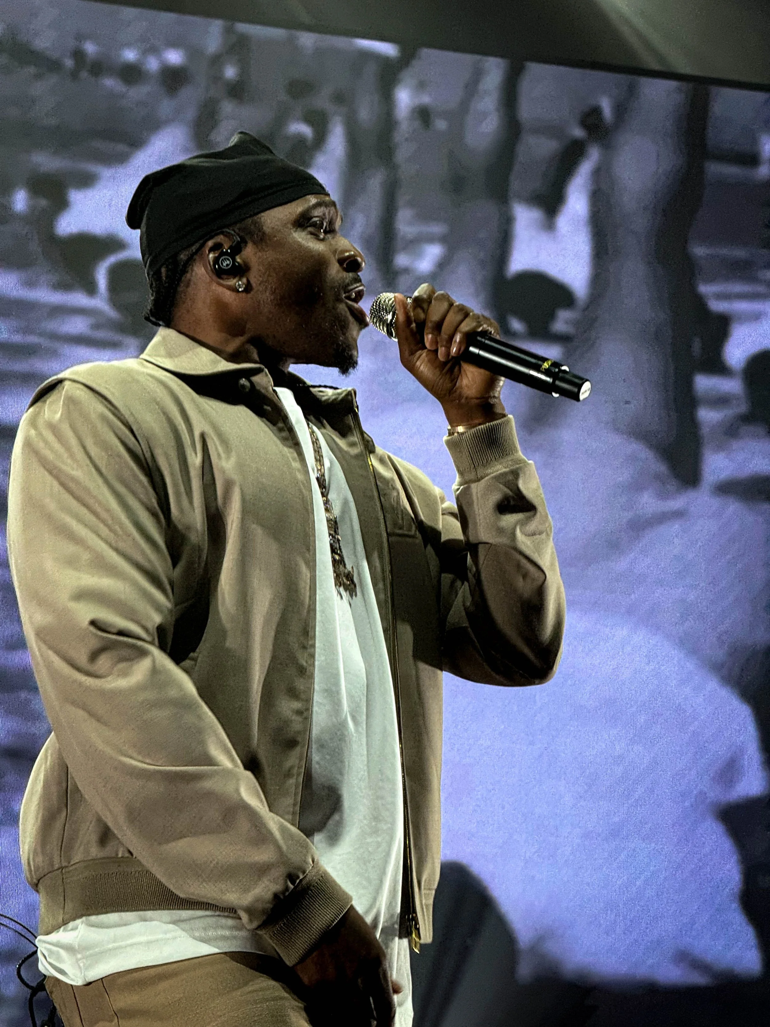 A man singing into a microphone on stage, wearing a beige jacket, white shirt, and a black head covering, with a large black-and-white image projected in the background.