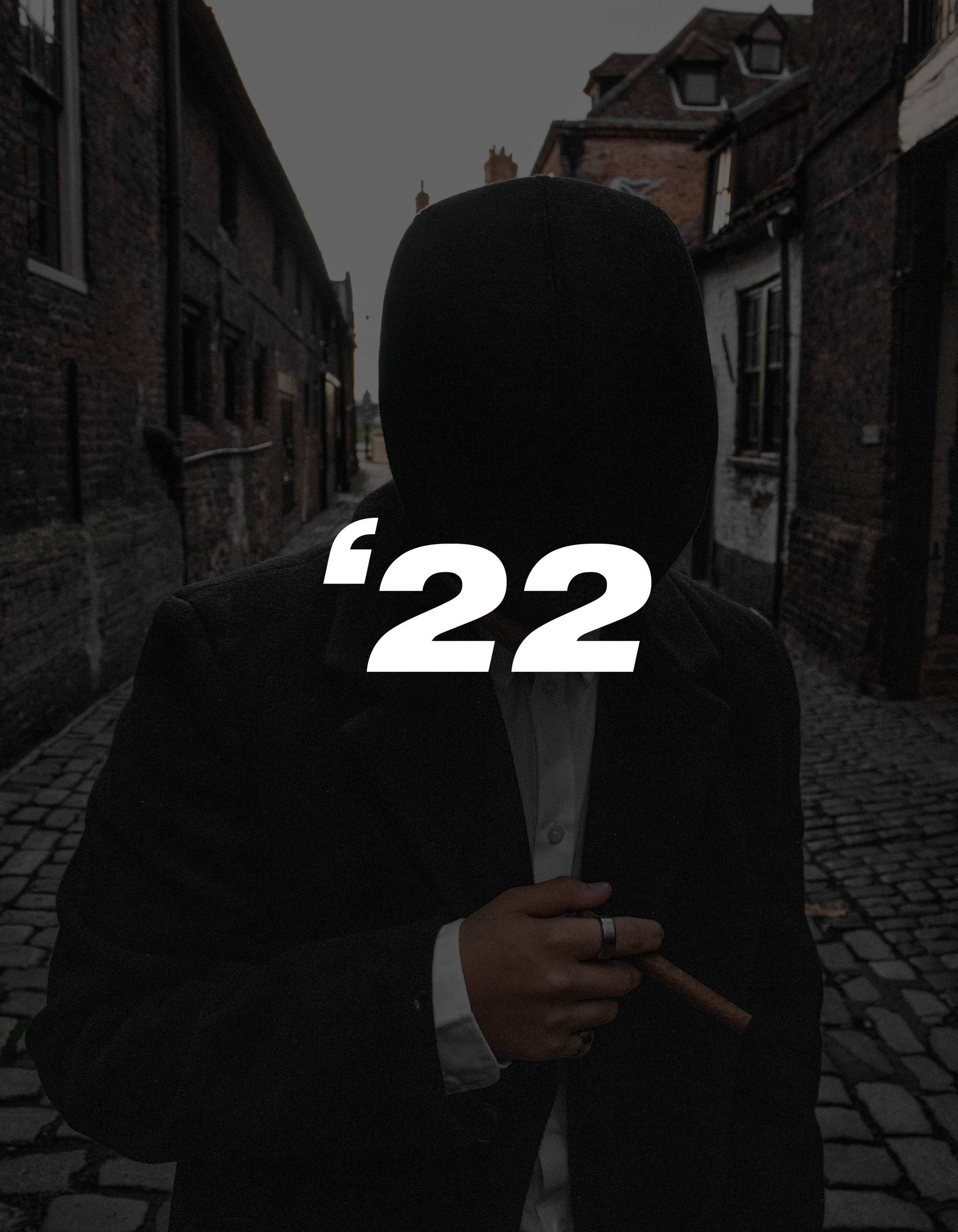 Person wearing a hoodie and suit jacket, holding a cigar, standing in an alleyway with cobblestone street and brick buildings in the background. The number '22' is overlaid on the image.
