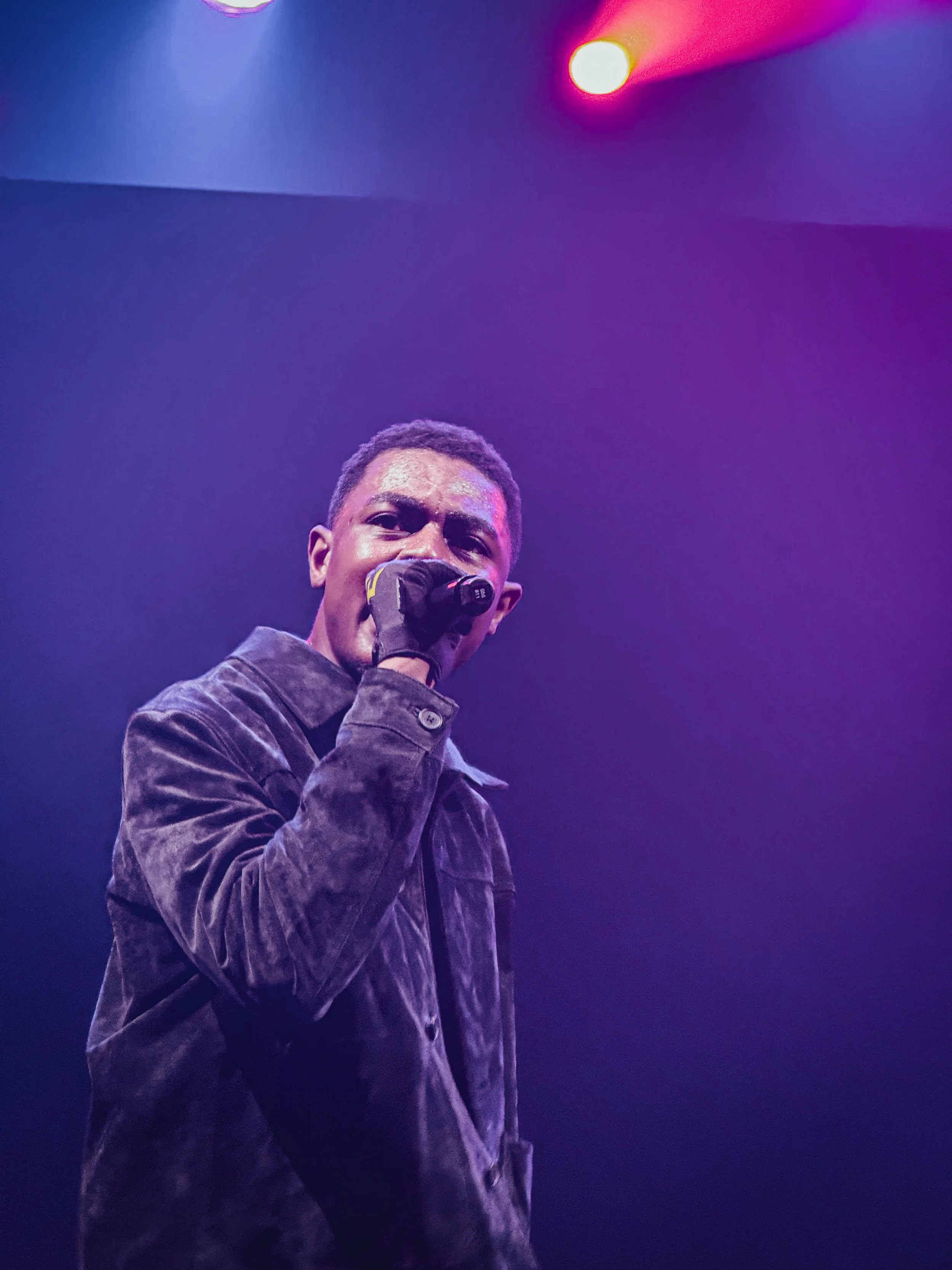 A man singing into a microphone on stage under colorful purple and yellow lighting.