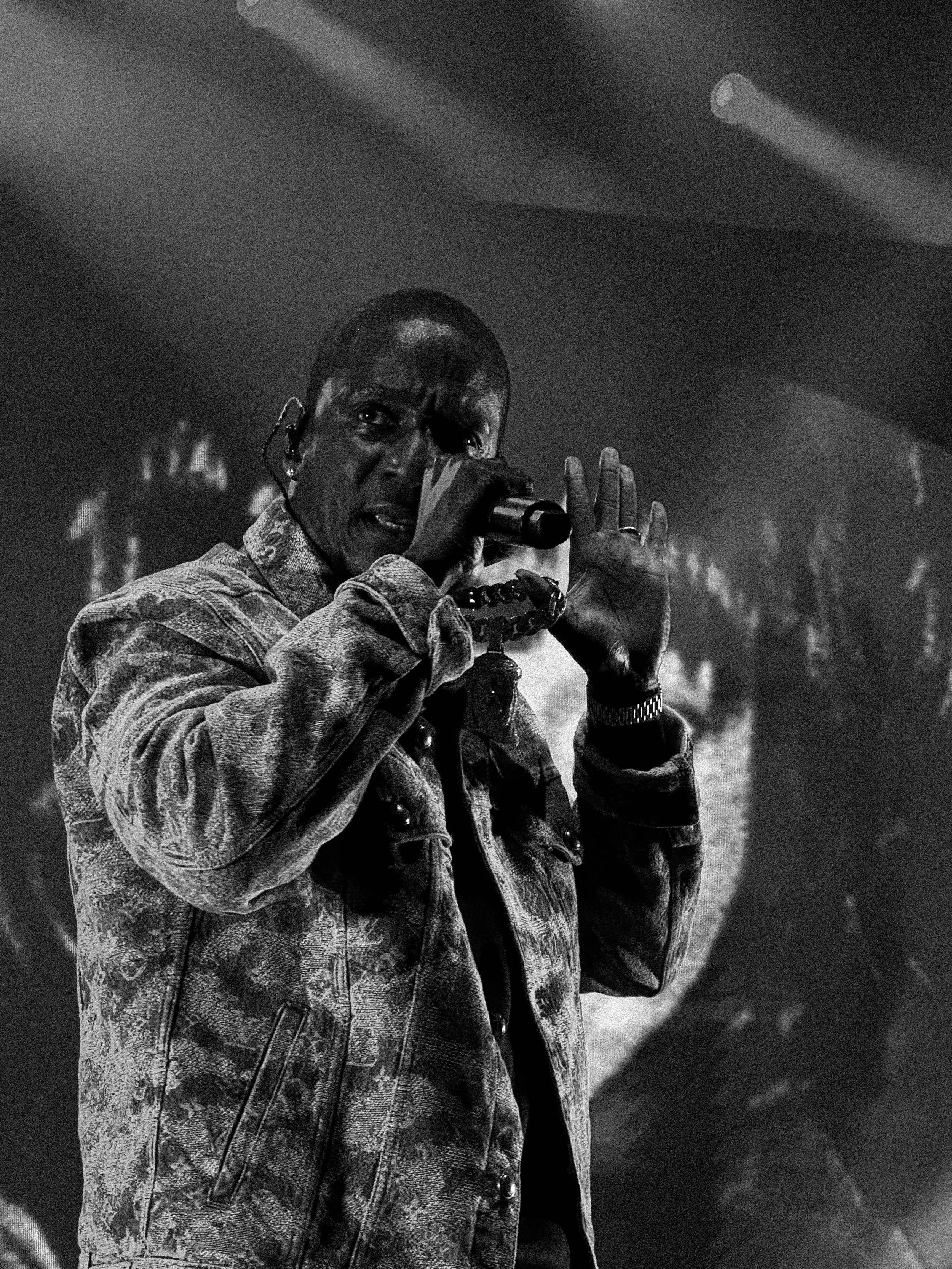 A man is singing or speaking into a microphone on stage, with a dark background. He is wearing a denim jacket and has short hair, with a serious expression.