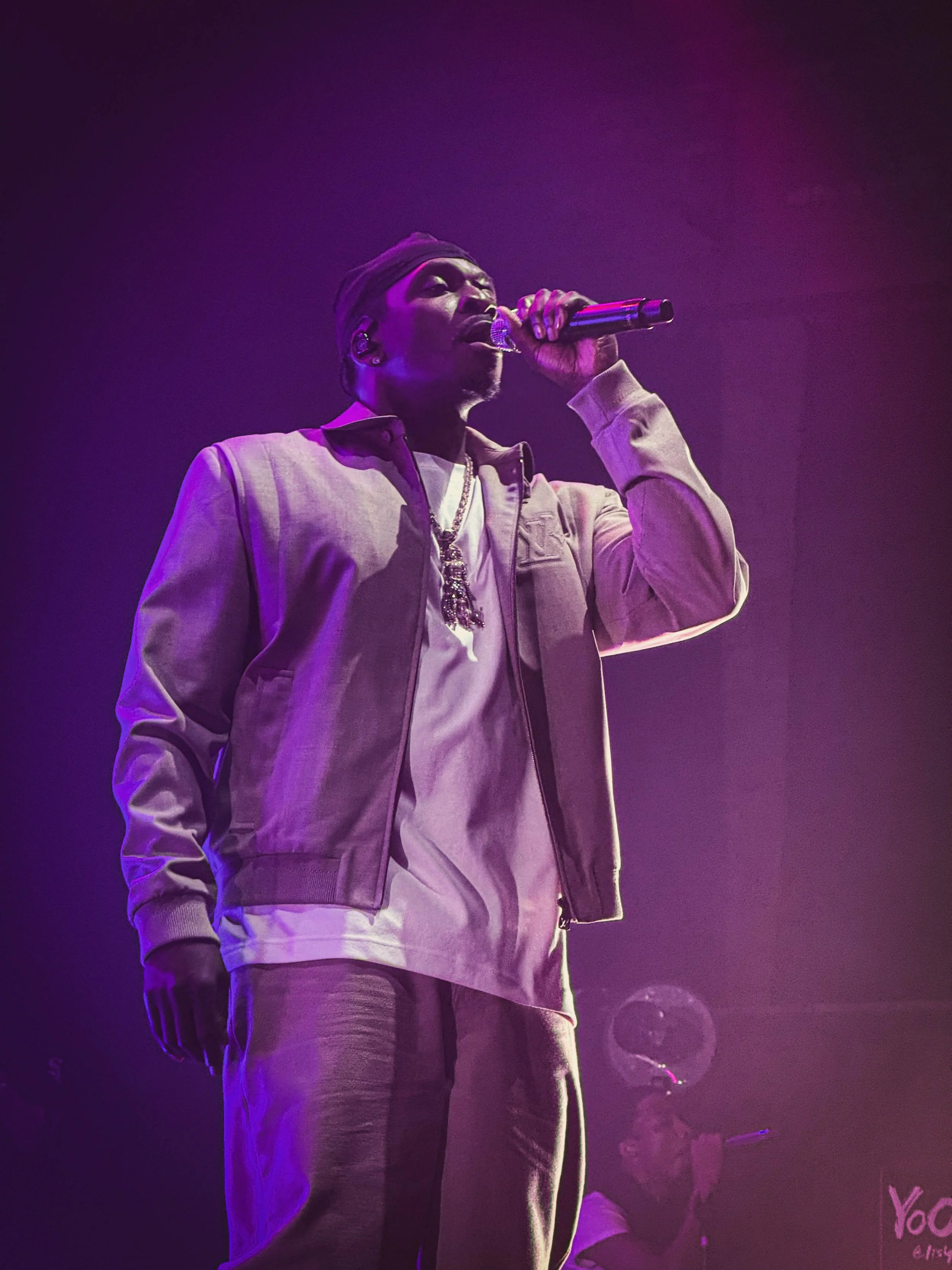A male singer on stage wearing a beige jacket, white shirt, and a necklace, holding a microphone and singing passionately, with purple stage lighting.