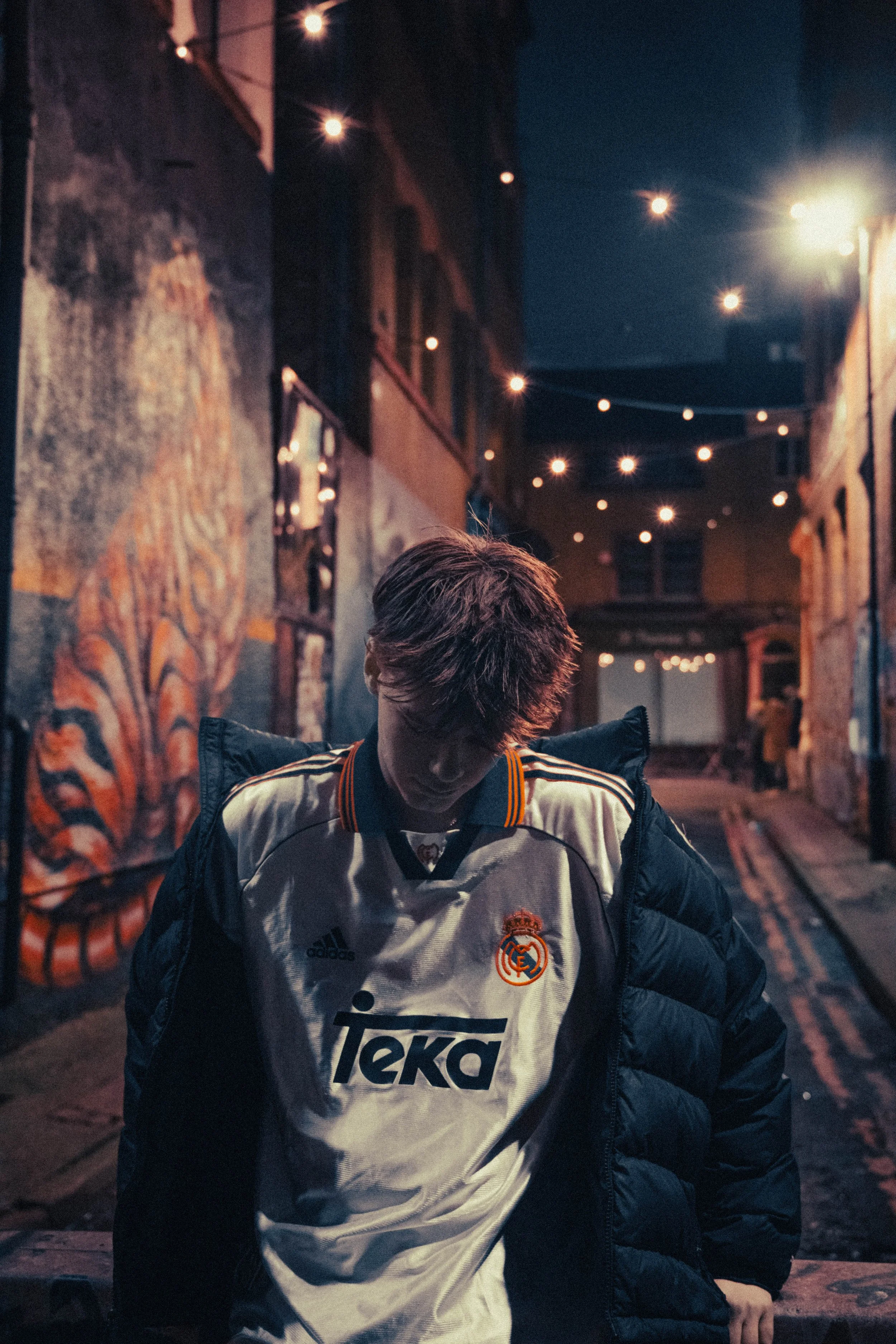 A young man in a white soccer jersey with the Real Madrid logo and the Teka sponsor, wearing a black puffer jacket, looking downward in a dimly lit urban alley with string lights overhead and graffiti on the walls.