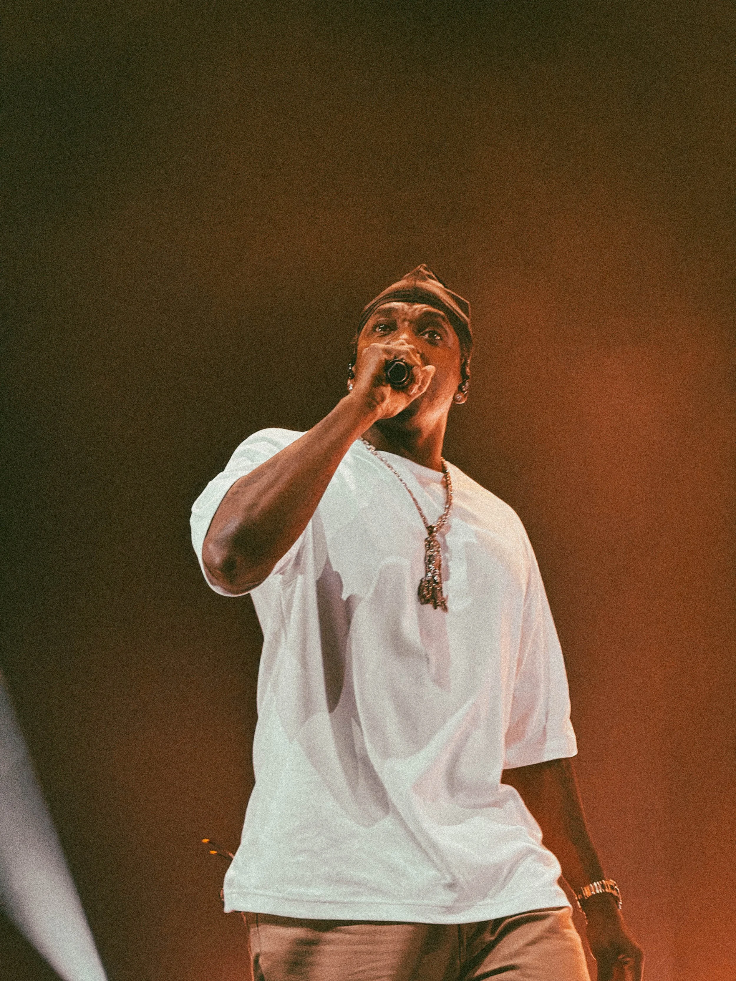 A man wearing a white shirt, gold chain, and a watch, holding a microphone, performing on stage.