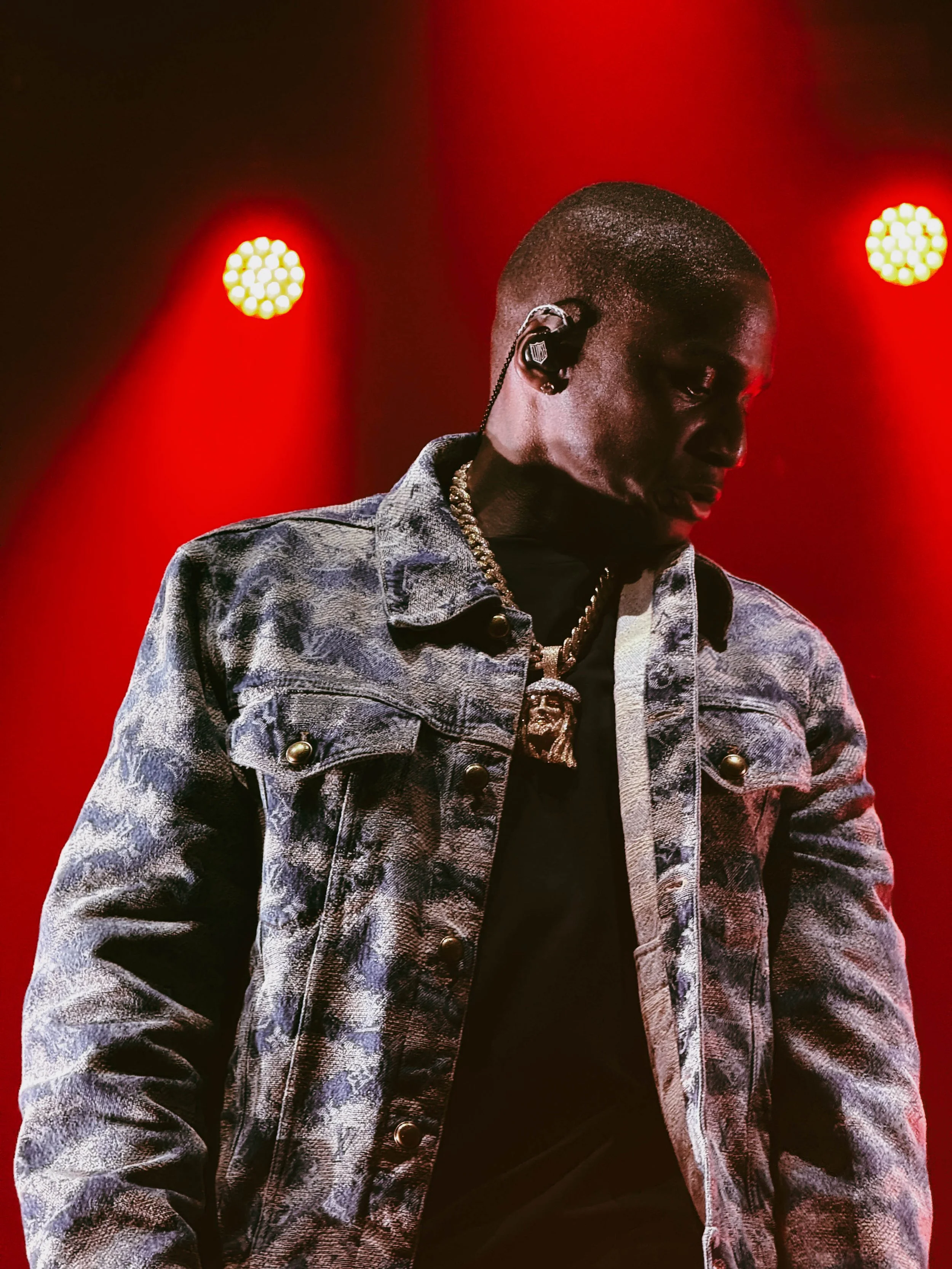 A male performer in a denim jacket and jewelry, wearing an in-ear monitor, standing on stage with red lighting and spotlights.