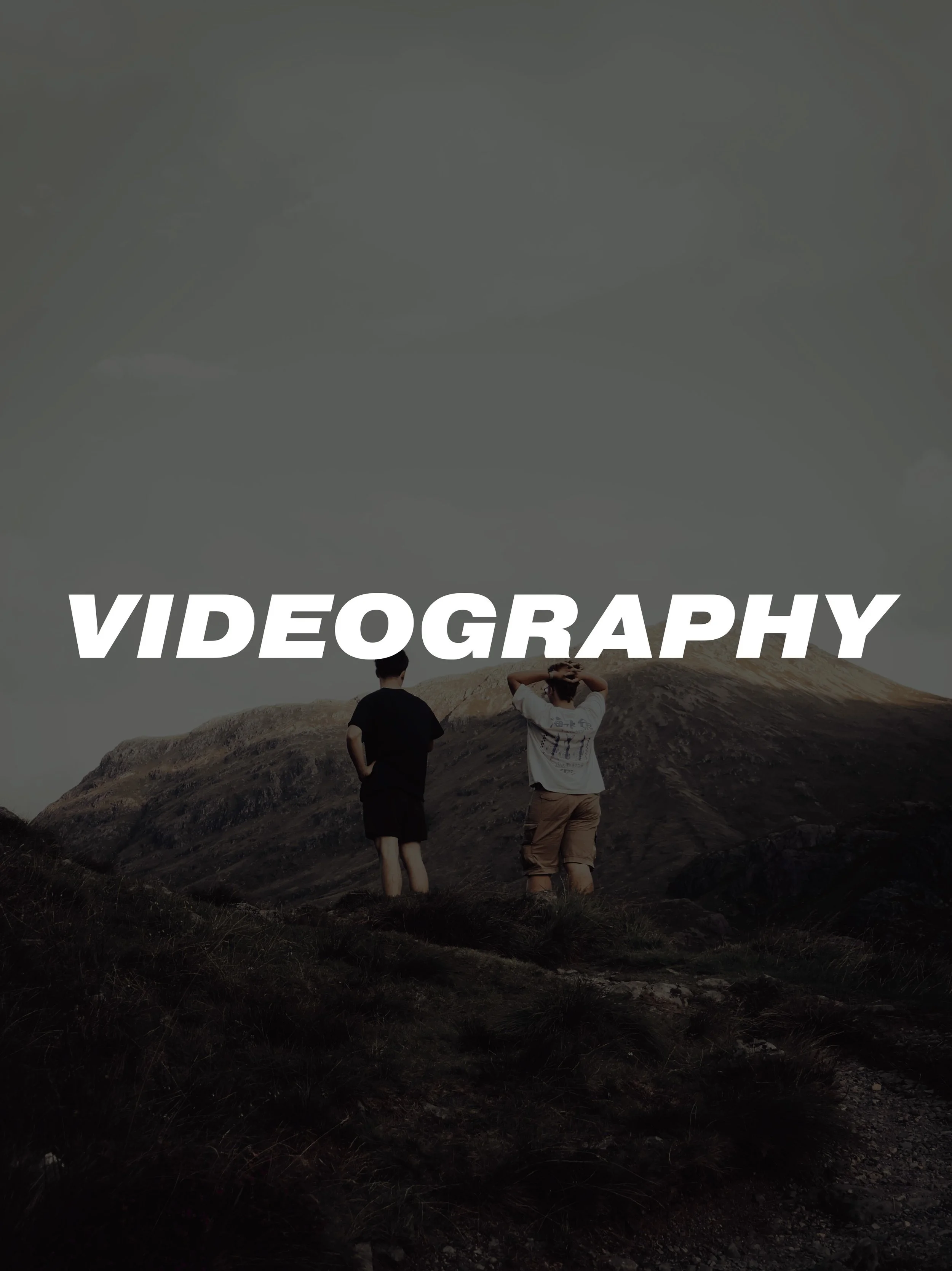 Two people standing on a mountain trail with mountains in the background, overlaid with the word 'VIDEOGRAPHY' in bold white letters.
