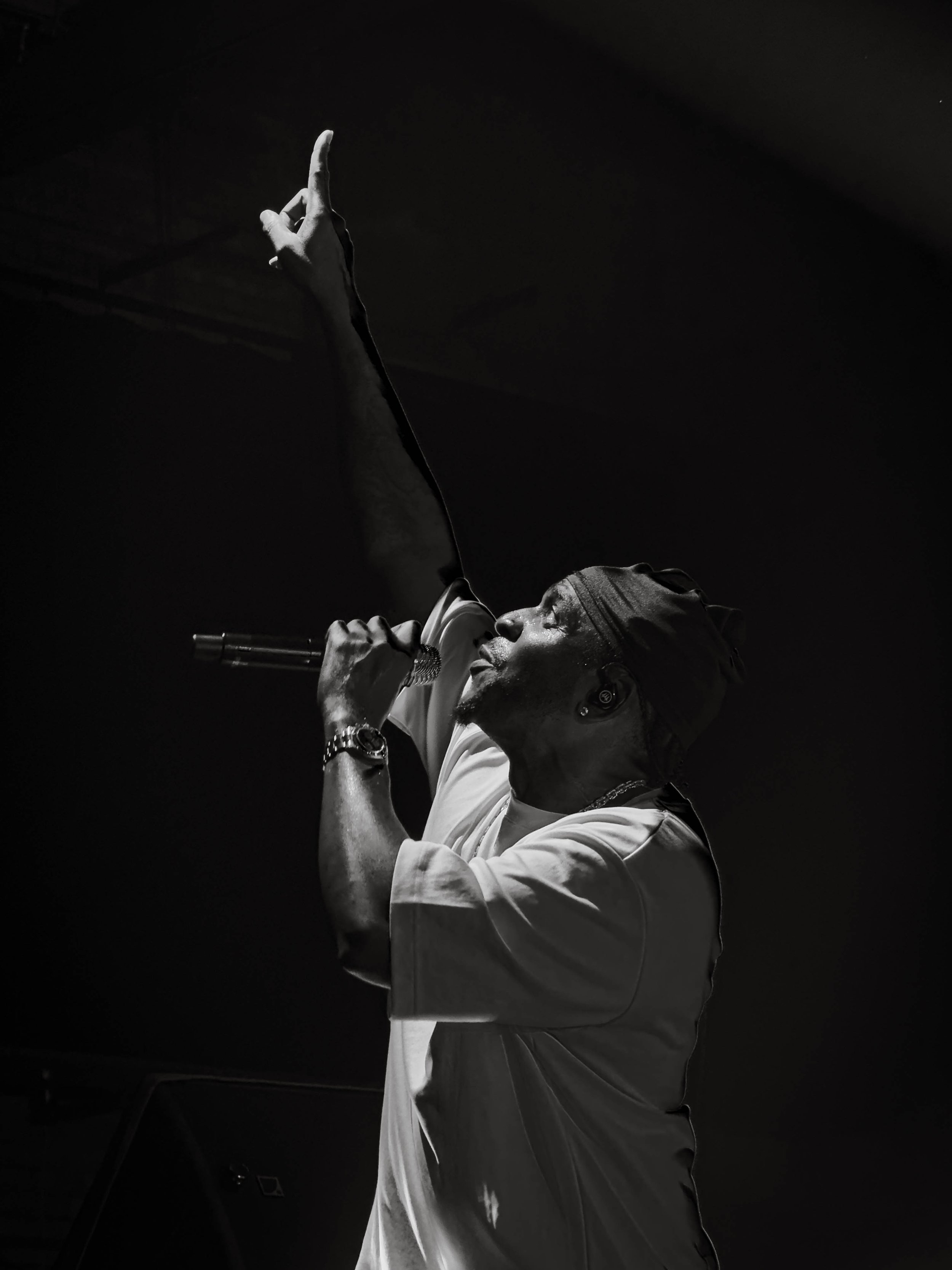 A man singing into a microphone, reaching upward with one hand, wearing a head covering and wristwatch, depicted in black and white.