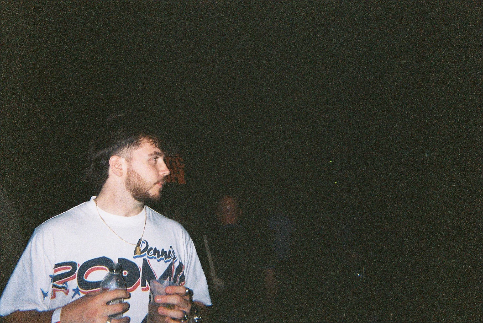 A young man with dark hair and a beard wearing a white T-shirt with red, blue, and black lettering and graphics, holding a drink in his hand at a dark indoor event.