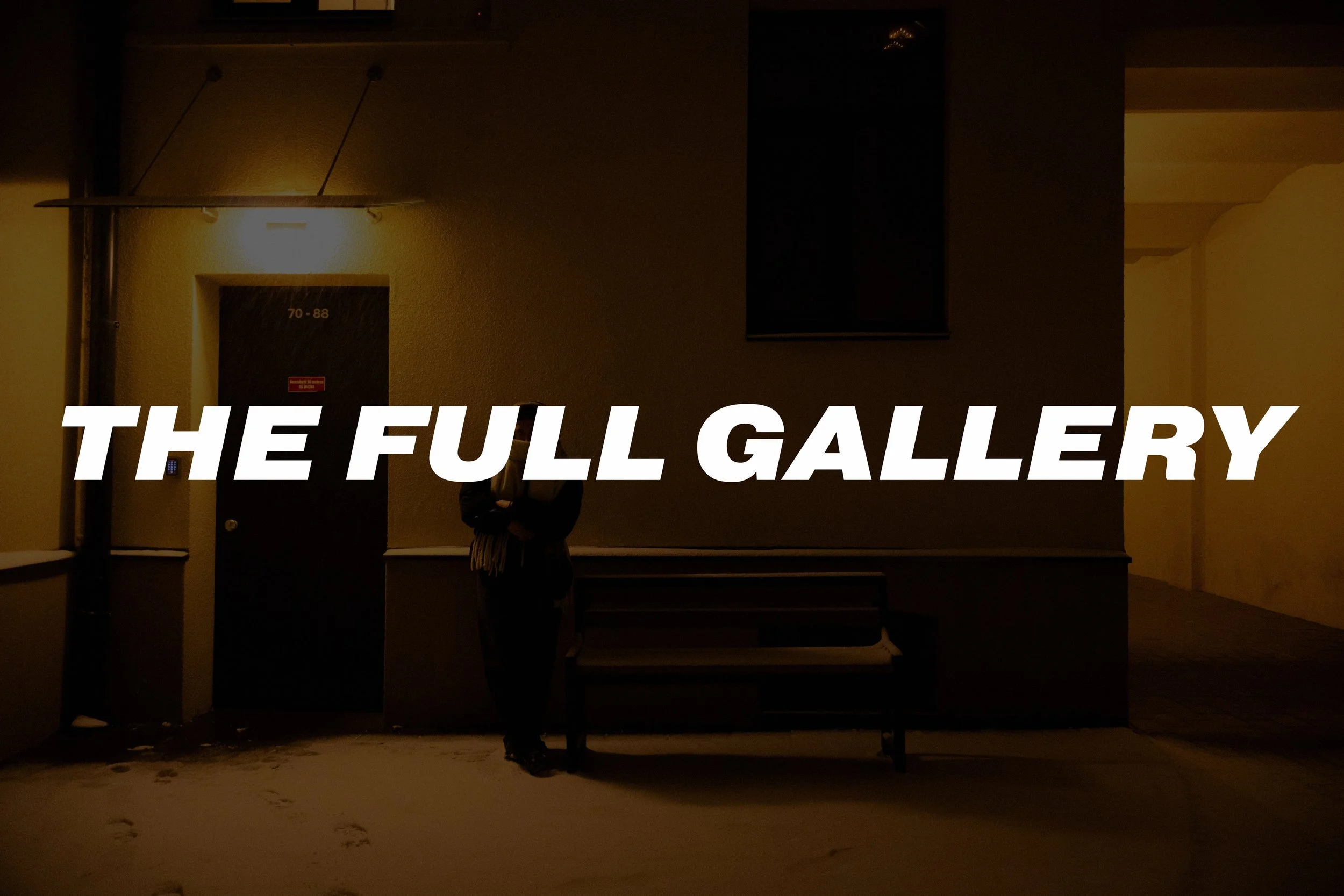 A person standing outside a building at night, illuminated by a yellow light, with a bench nearby and snow on the ground. The words 'THE FULL GALLERY' are overlaid in bold white text.