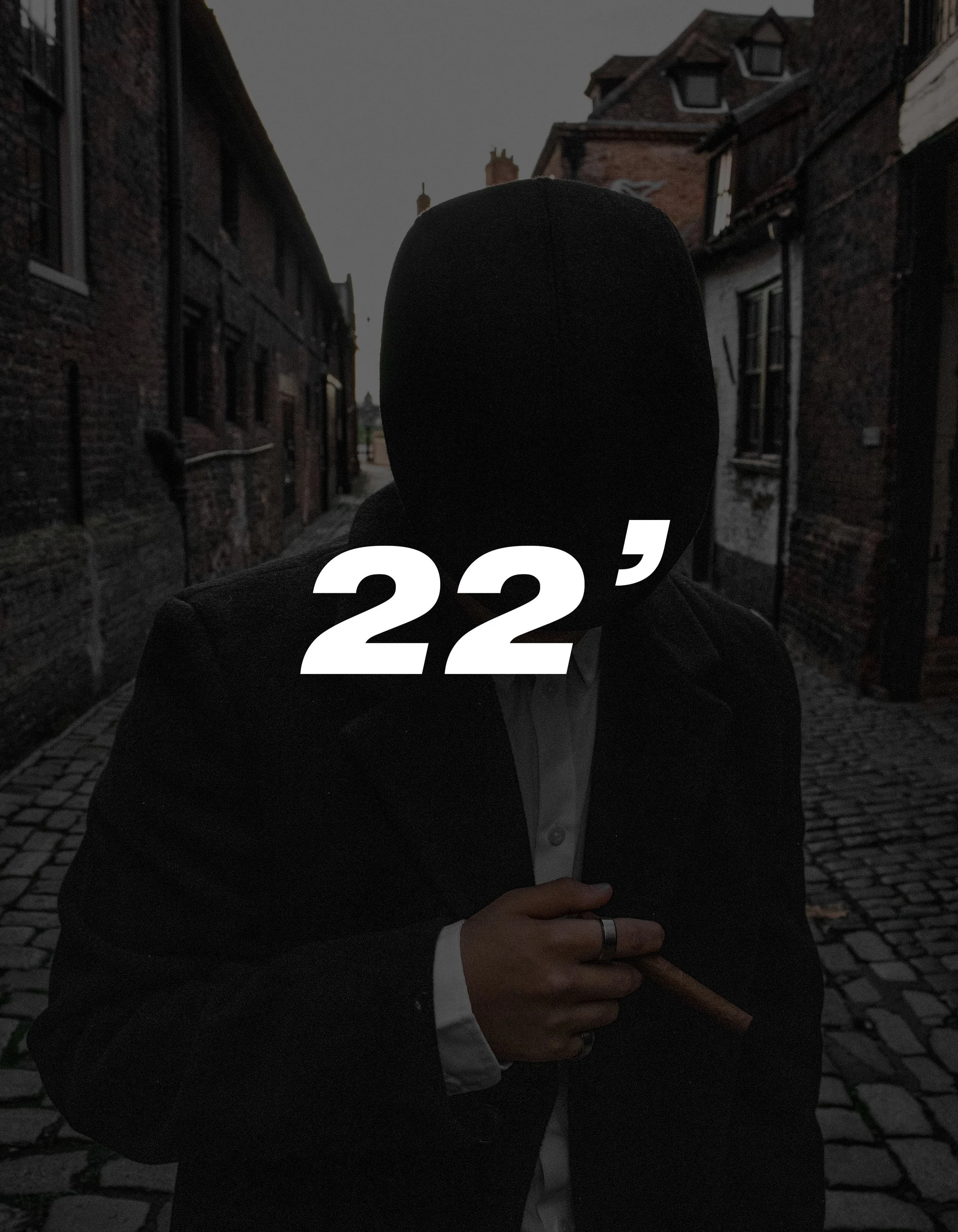 A person wearing a hoodie and suit jacket standing on a cobblestone street in a narrow alley, holding a cigar, with large white '22' overlayed on the image.