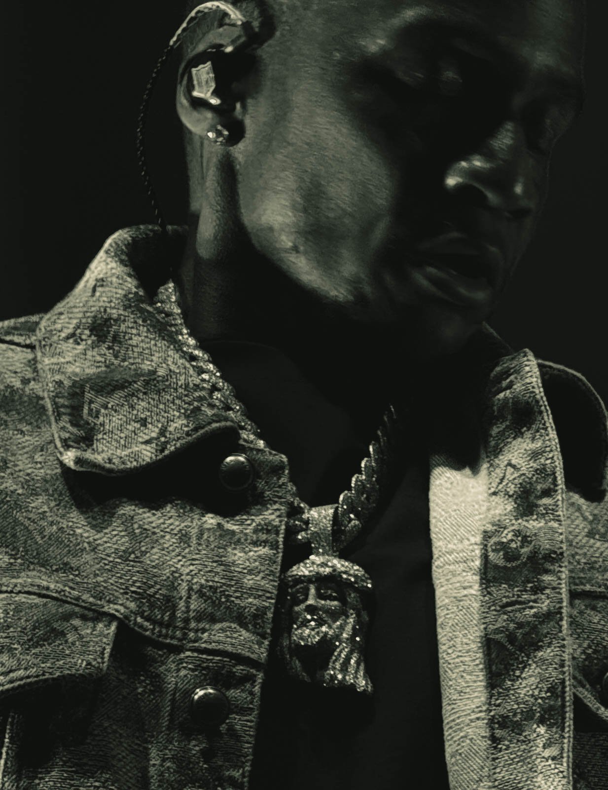 Close-up of a person wearing a denim jacket, large necklace with a face pendant, and earrings, in black and white.