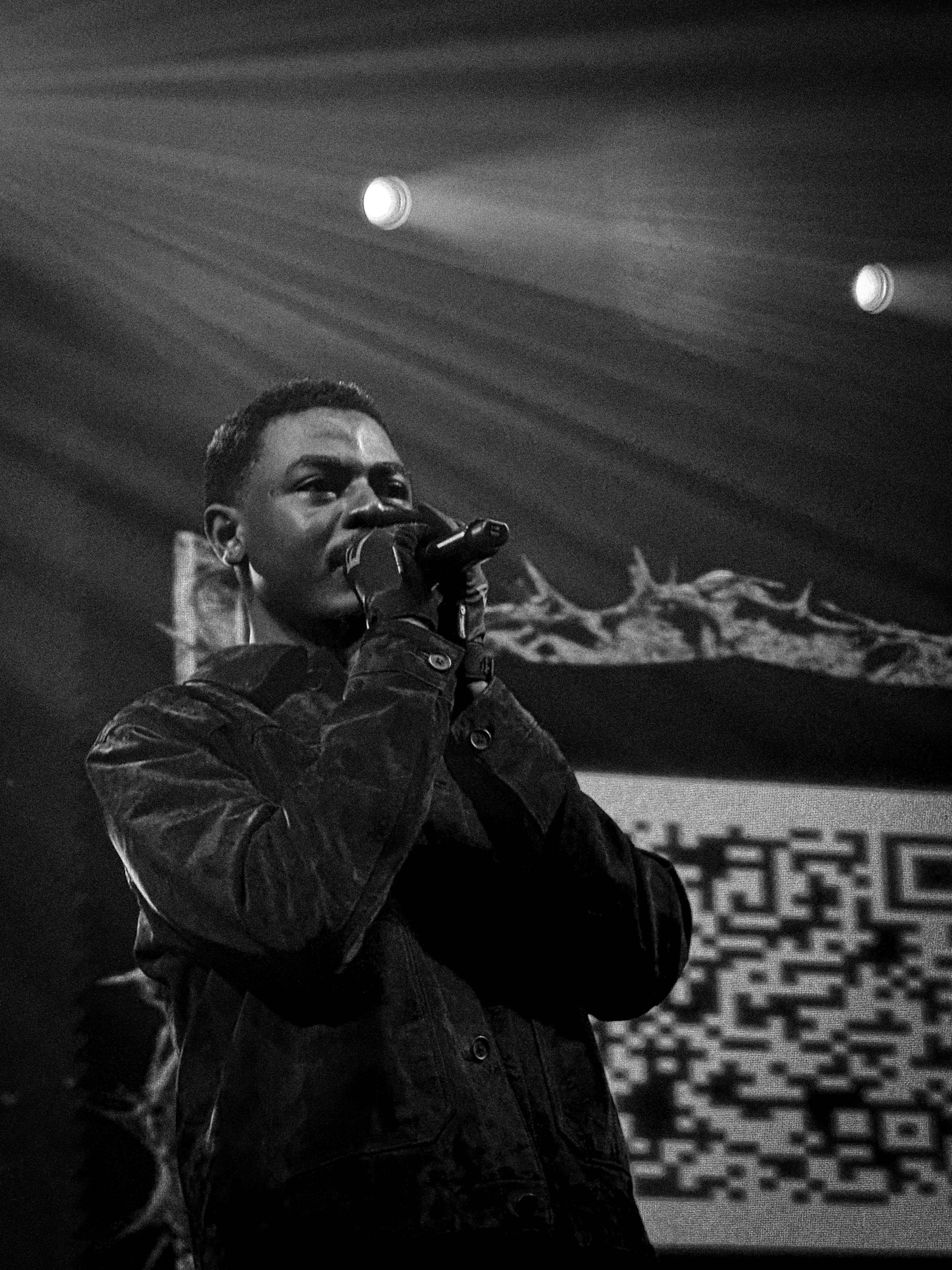A person singing into a microphone on stage with spotlights overhead and a digital QR code display in the background.