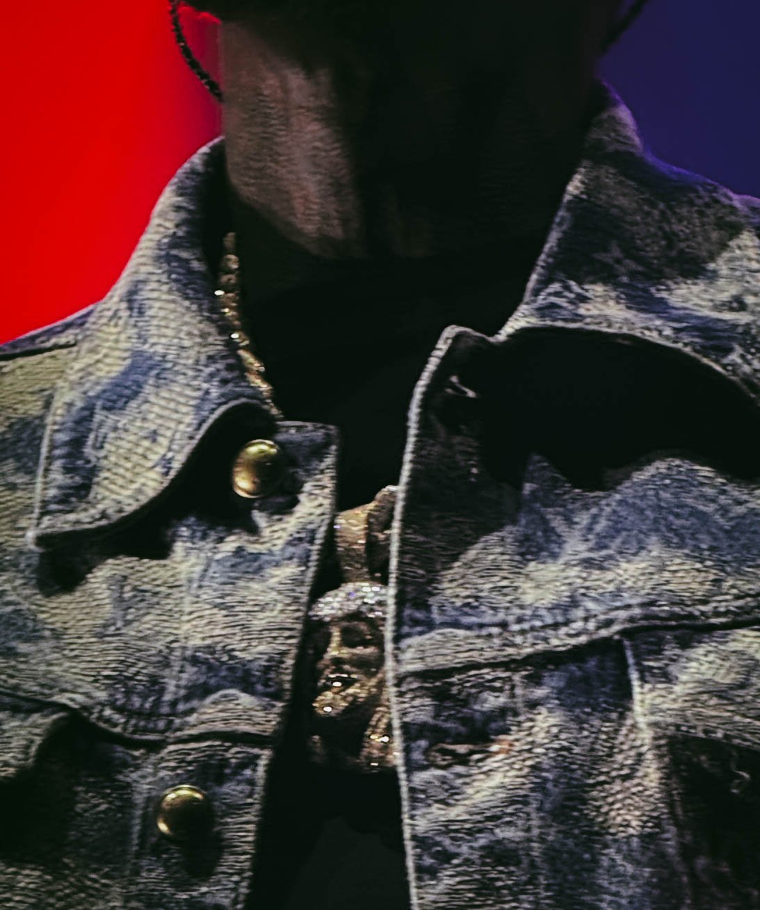 Close-up of a person wearing a denim jacket with a black turtleneck underneath, with a dark and colorful background.