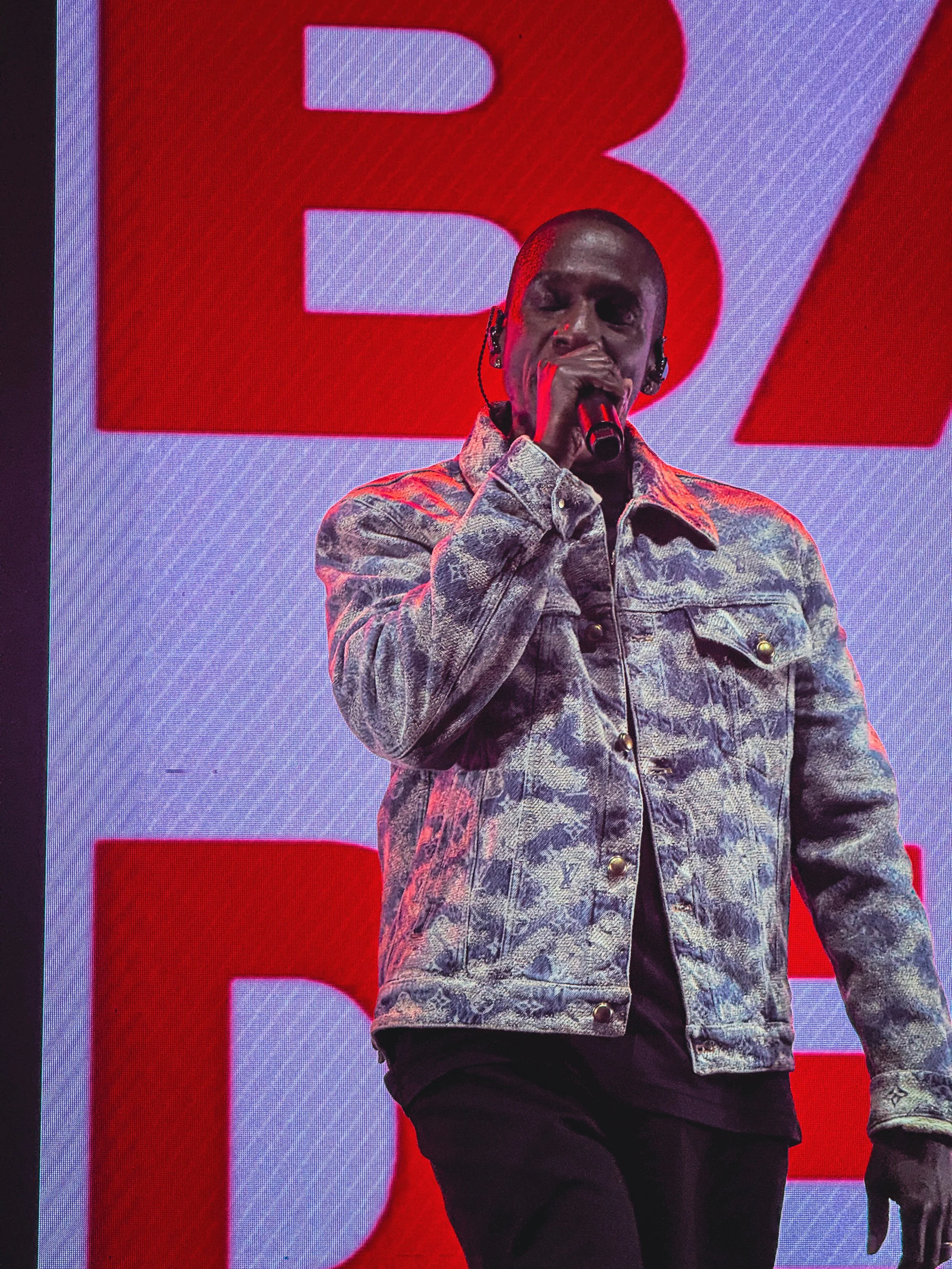 A performer on stage singing into a microphone, wearing a denim jacket with a patterned design, in front of a large screen displaying a red and white logo or text.