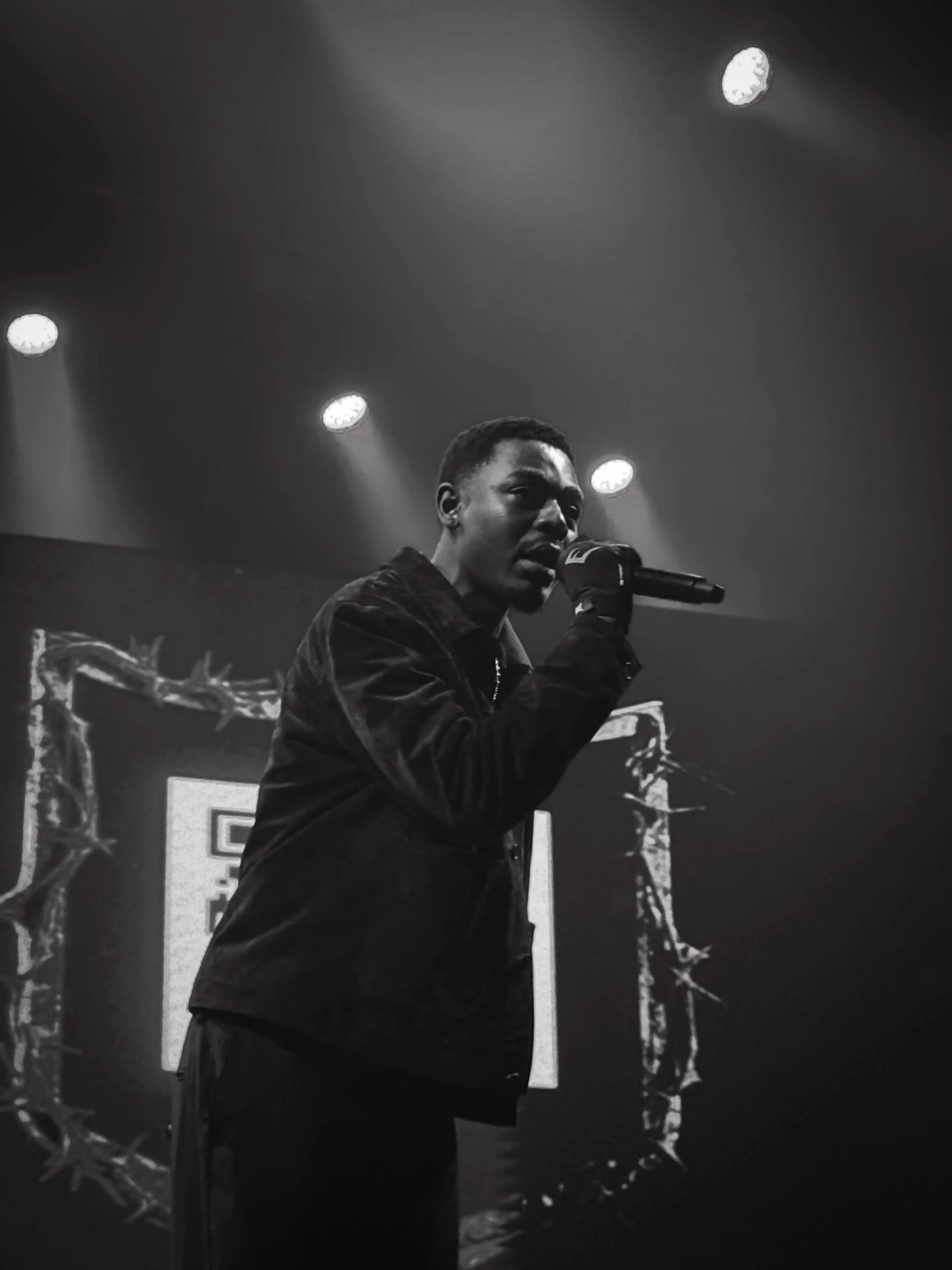A black and white photo of a male singer holding a microphone on stage, with stage lights and a decorative frame in the background.