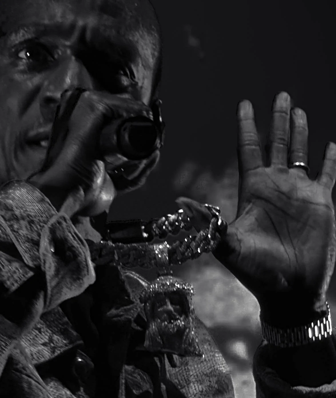 Close-up black-and-white photo of a man holding a camera up to his eye, wearing a watch, rings, and layered jewelry, with his left hand raised in a waving gesture.
