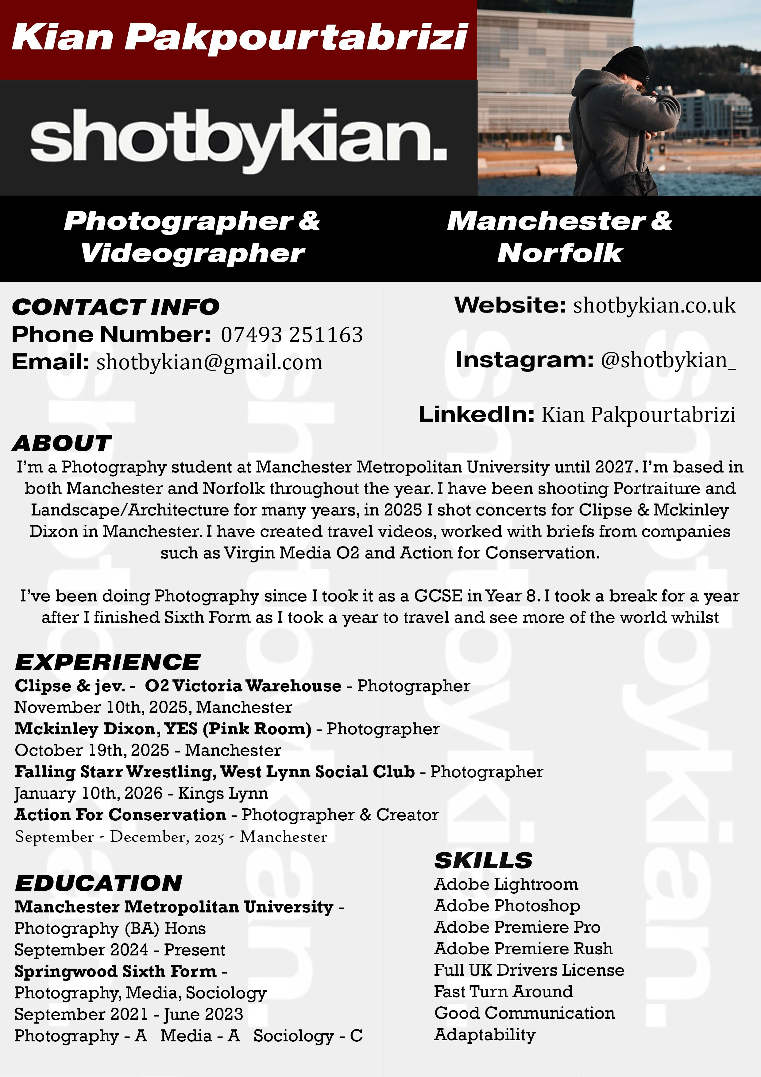 A man standing outdoors on a waterfront, wearing a black hoodie and beanie, with buildings and water in the background.