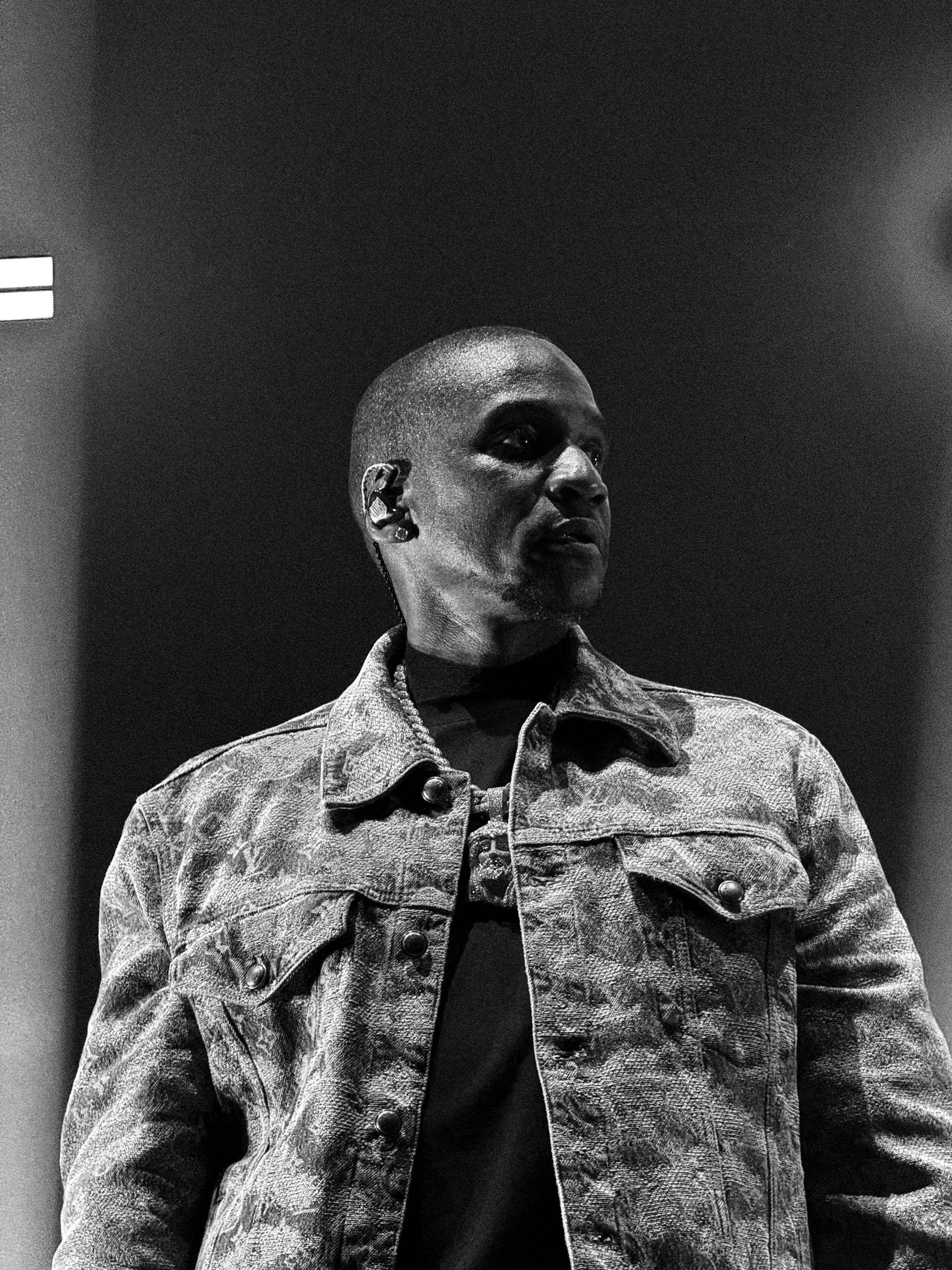 A person with a short hairstyle and wearing a denim jacket, standing in a dimly lit environment, looking to the side.