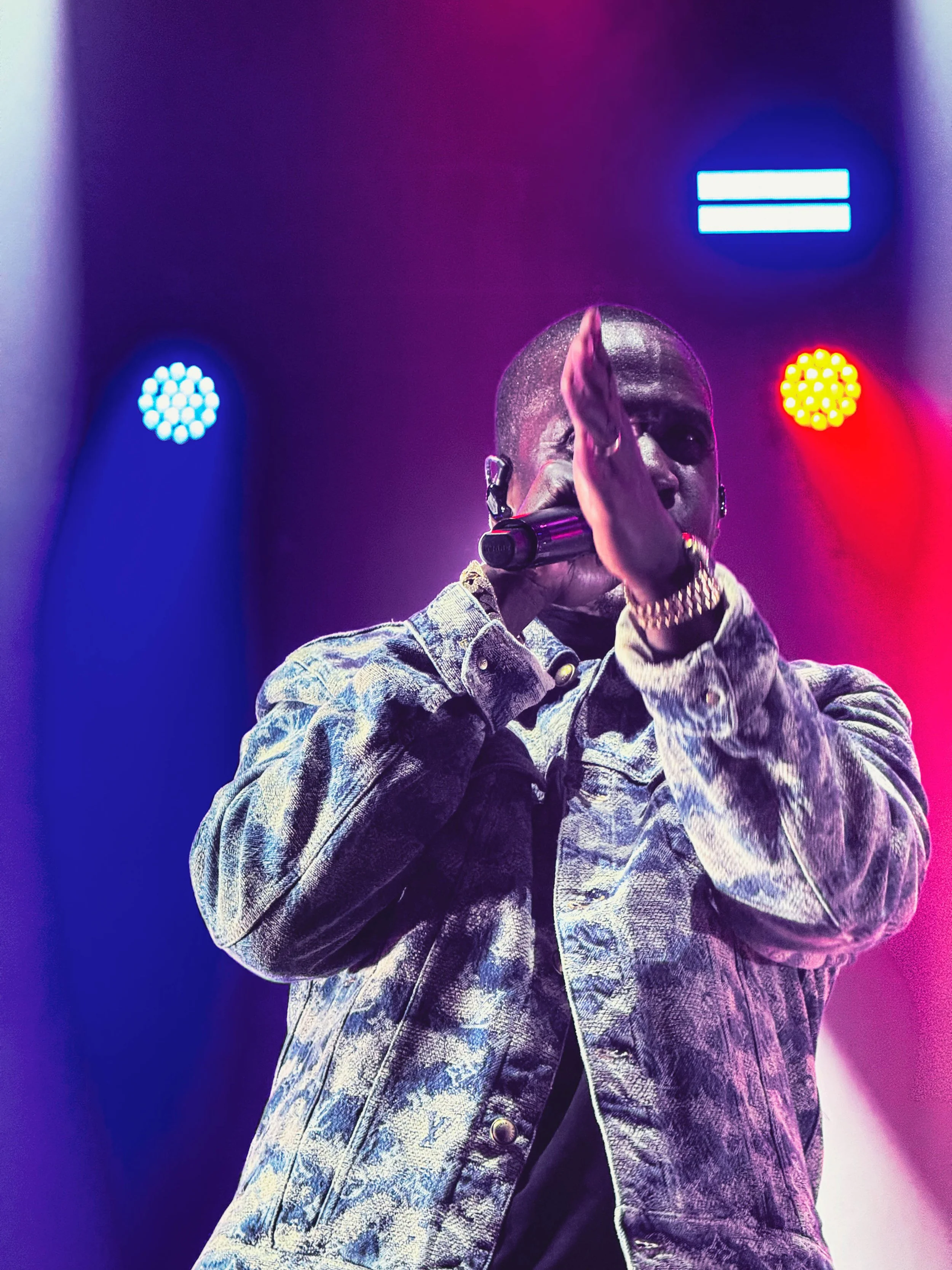 A man holding a microphone and gesturing with his hand during a performance on stage, with colorful blue, red, and purple lights in the background.