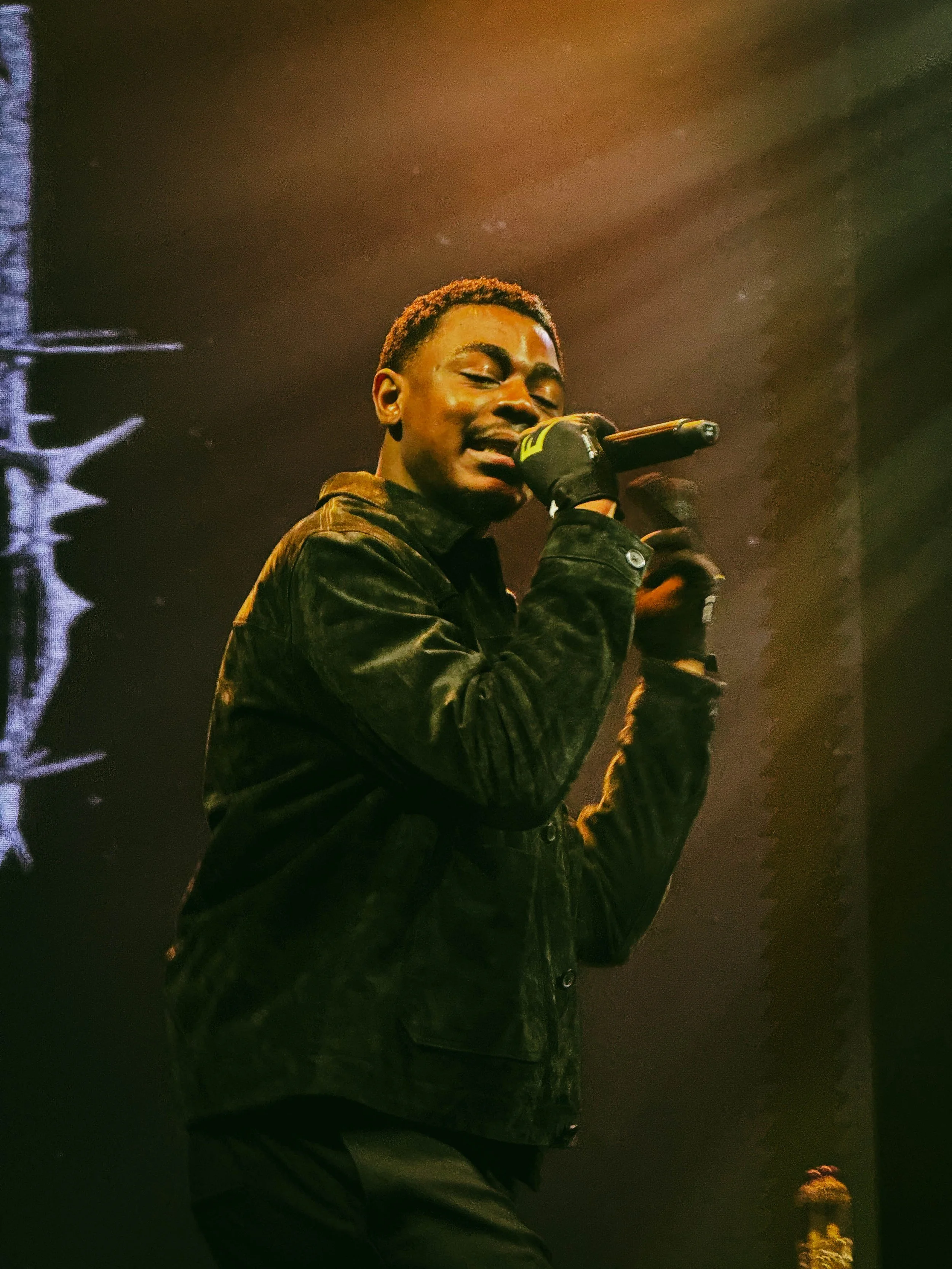 A young man singing into a microphone on stage, wearing a black jacket and gloves, with stage lights in background.