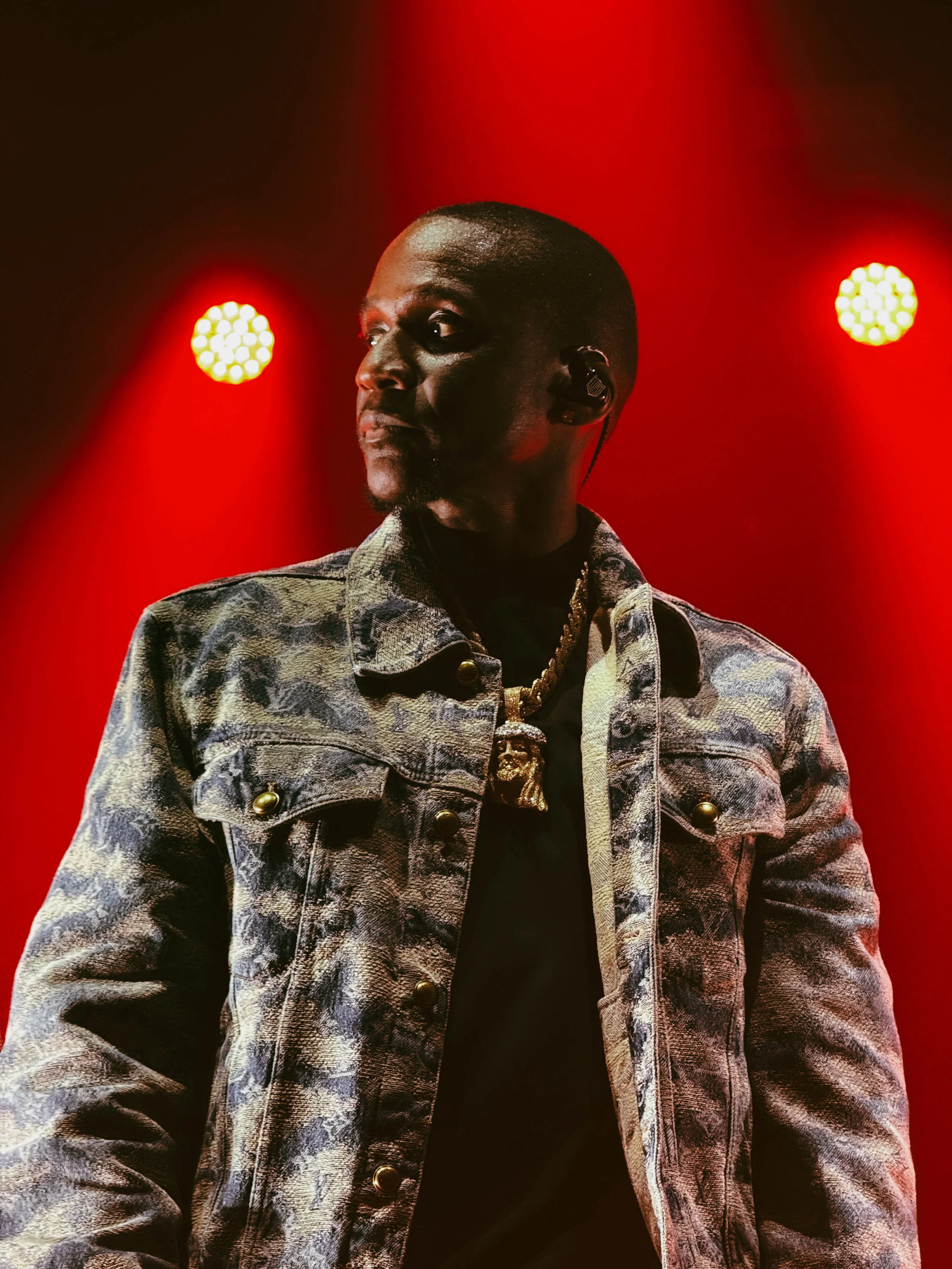A male performer on stage with red lighting, wearing a patterned denim jacket, a black shirt, and a gold chain.