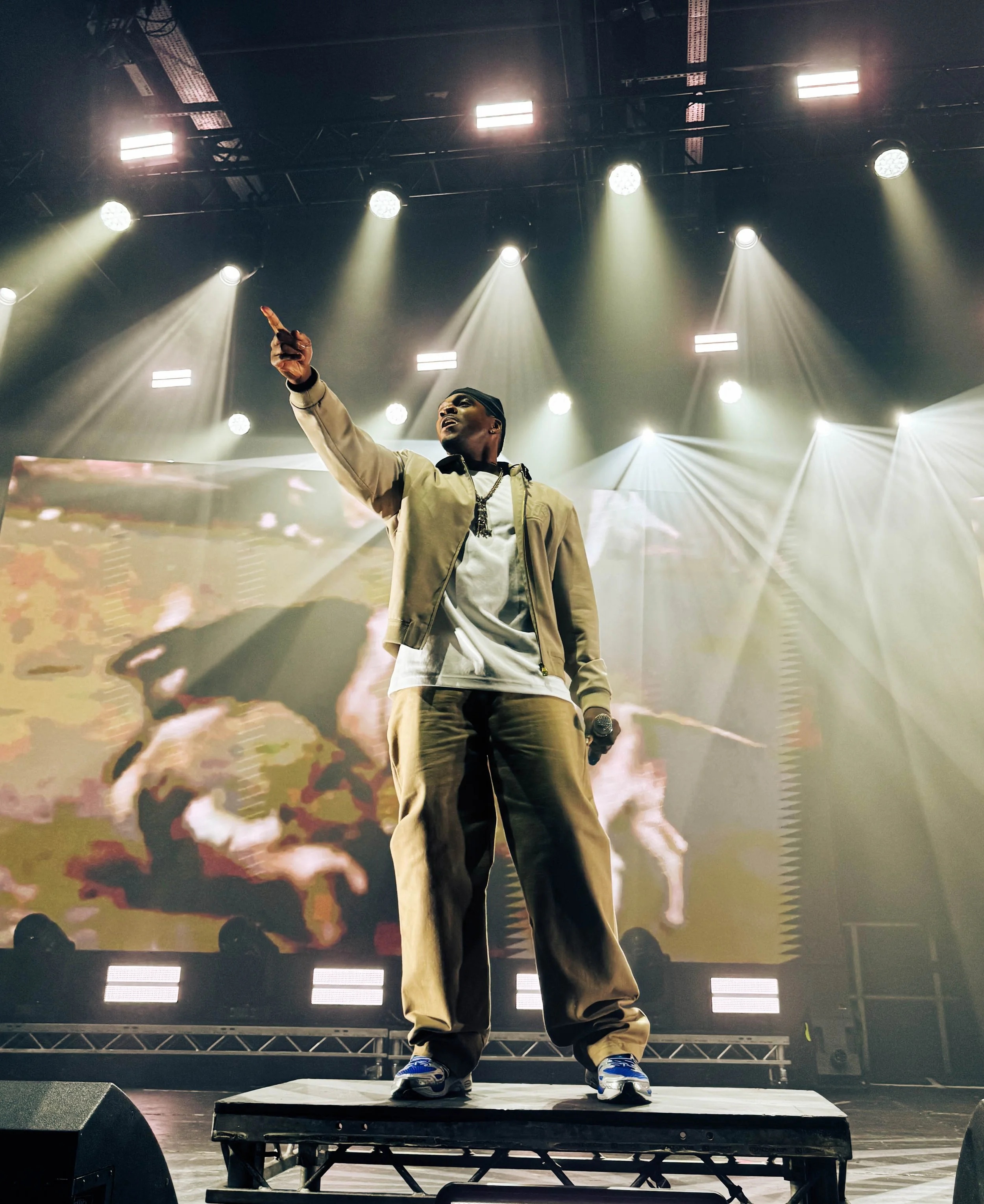 A male performer on stage under bright lights, pointing upward with one hand, with a large screen displaying abstract visuals behind him.
