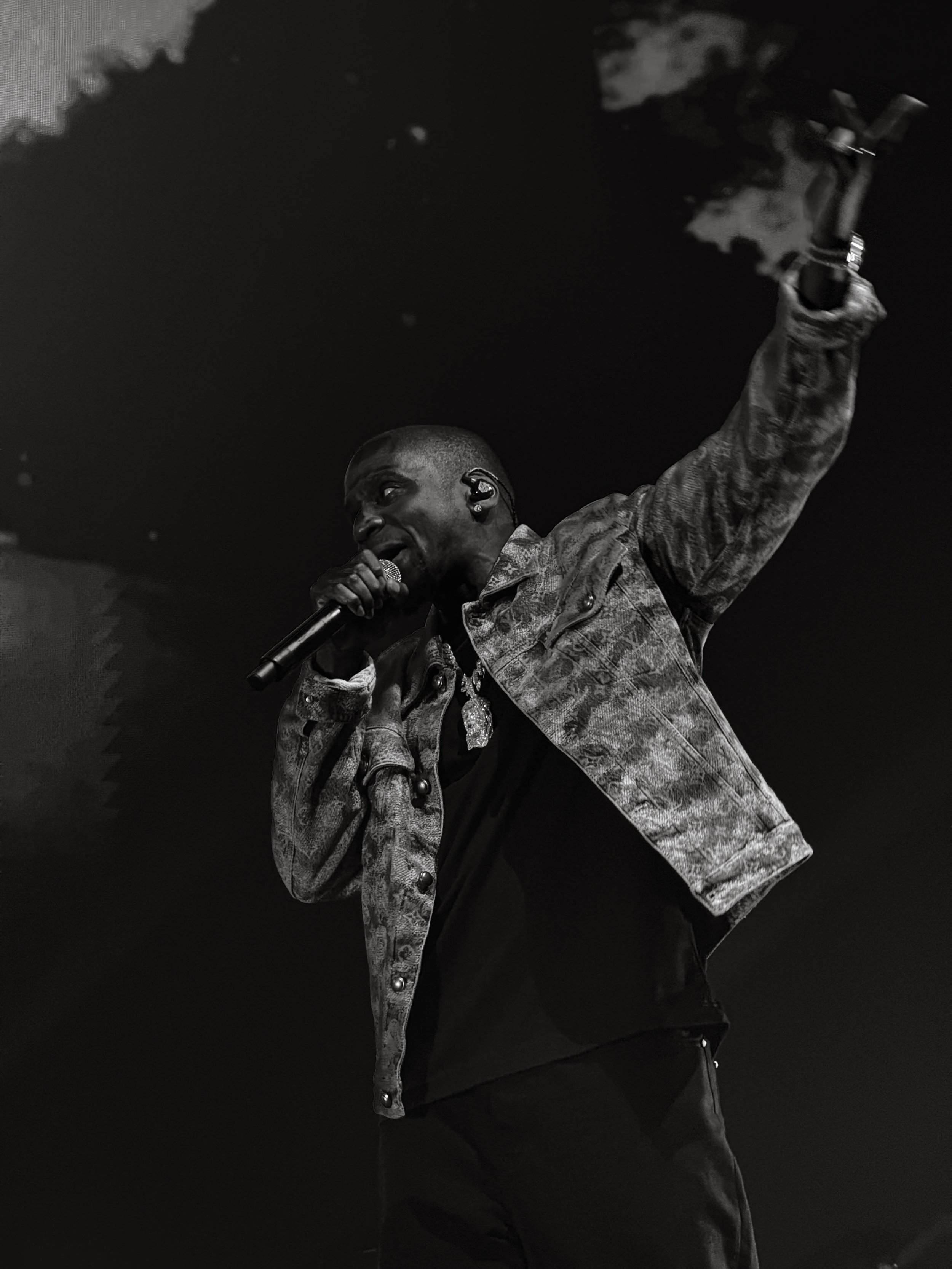 A male performer is singing into a microphone on stage, wearing a patterned jacket and dark clothing, with one arm raised.