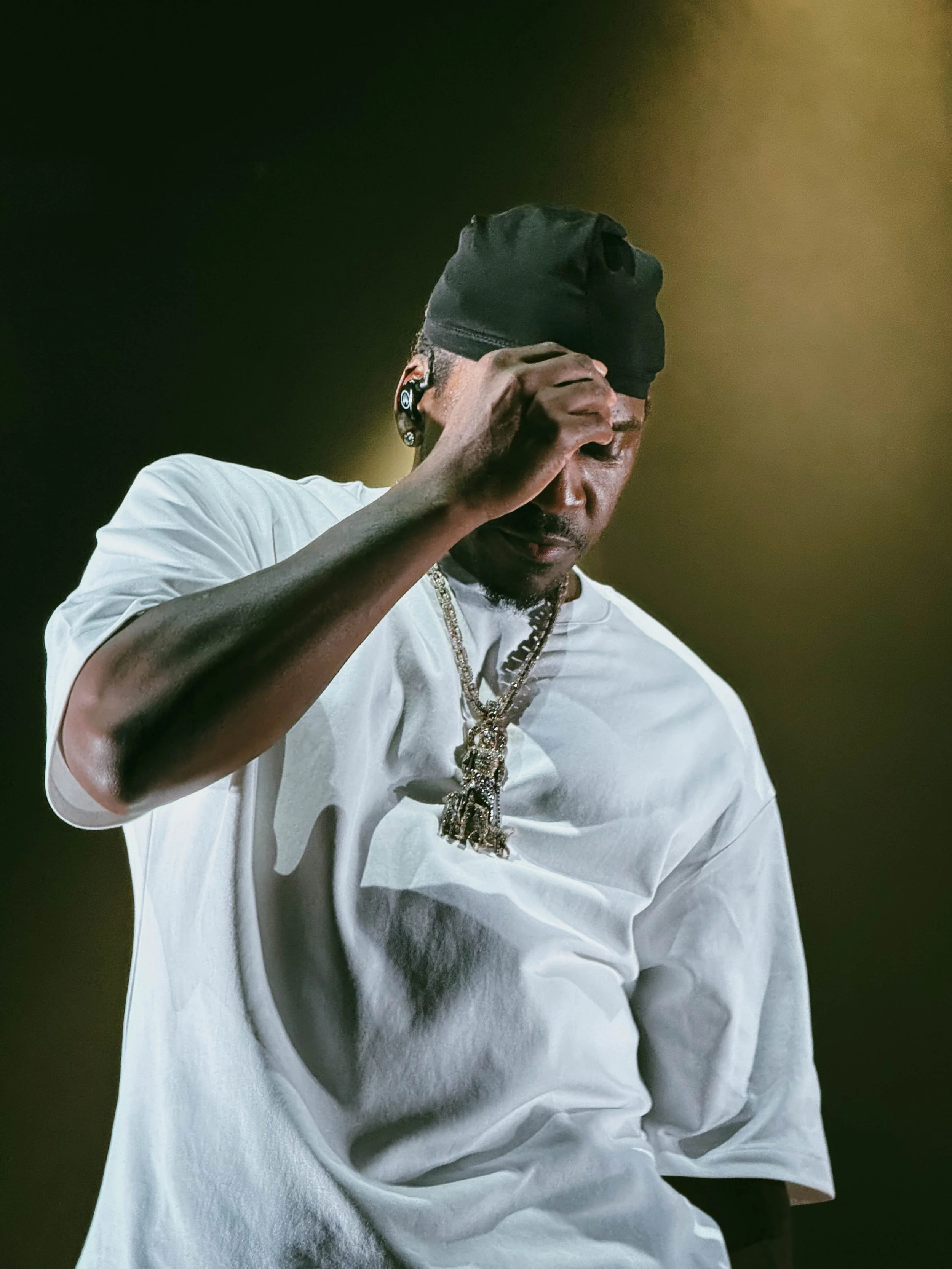 A man wearing a black beanie, a white T-shirt, and jewelry, holding his hand up to his face during a performance or concert.