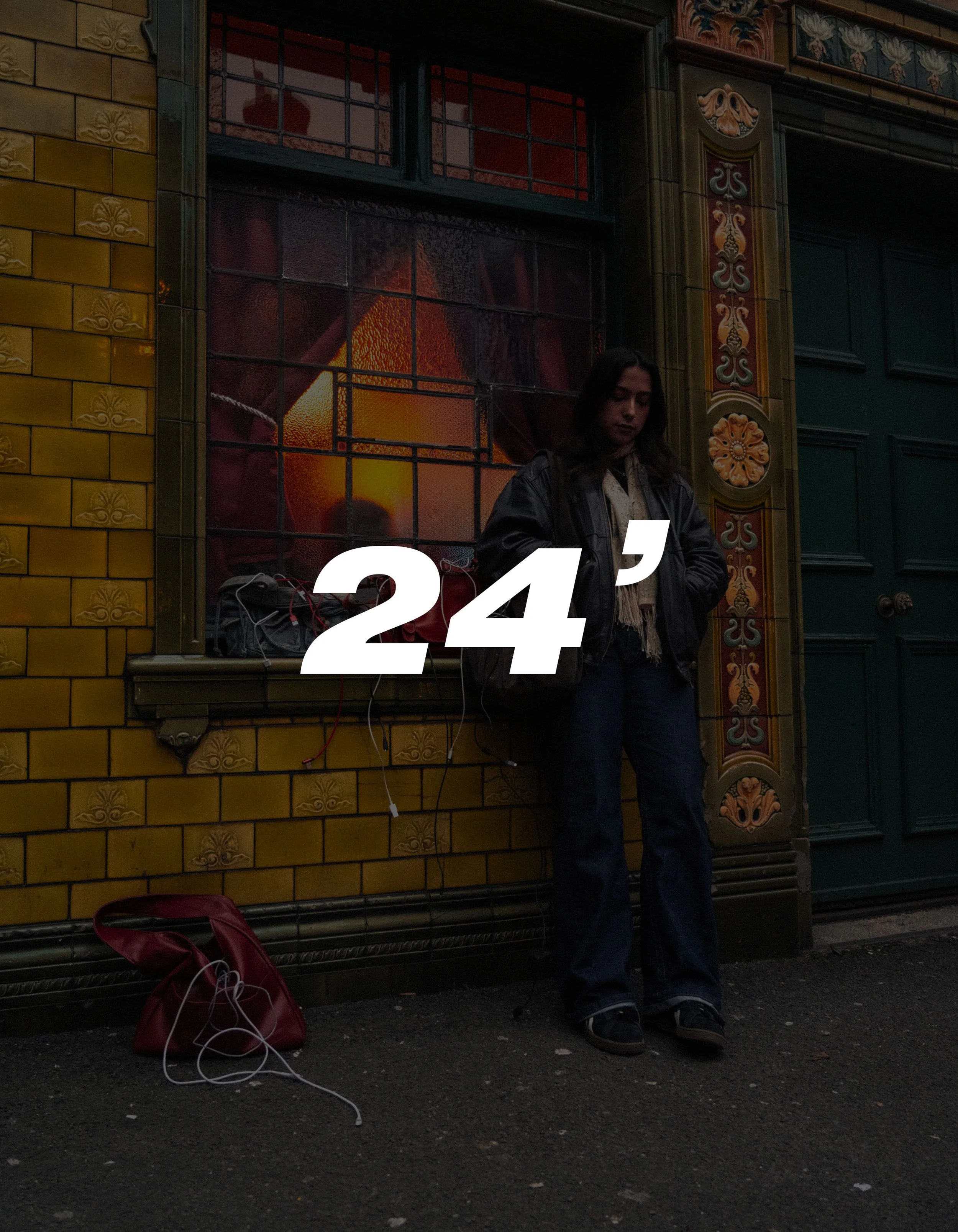 A woman with long dark hair standing against a yellow tiled building with ornate green door and window frame, next to a red bag on the ground, with a window display showing a sunset scene in the background.